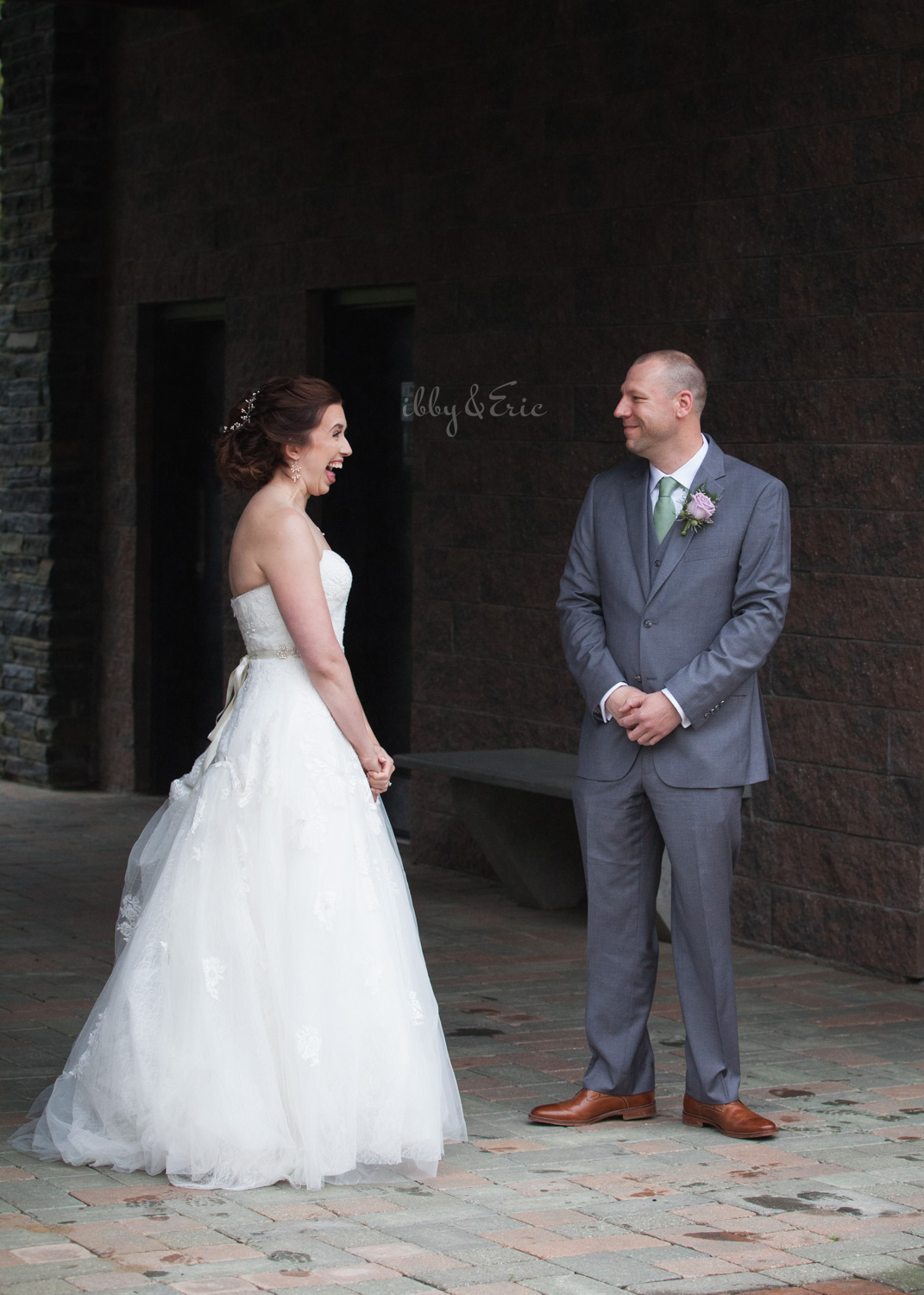 Groom in a gray suit smiles as he sees his bride in a lace wedding gown for the first time.
