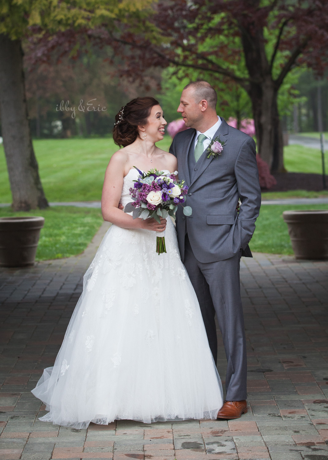 Bride and groom smile at each other while holding a purple and white bouquet.