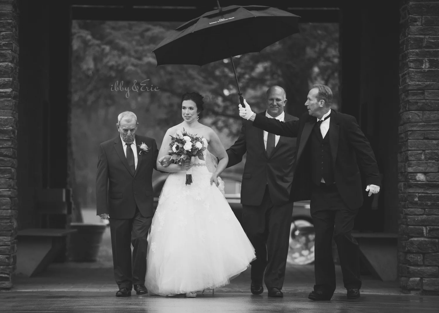 Driver holds a black umbrella over the bride, as she is escorted down the aisle by her dad and grandpa.