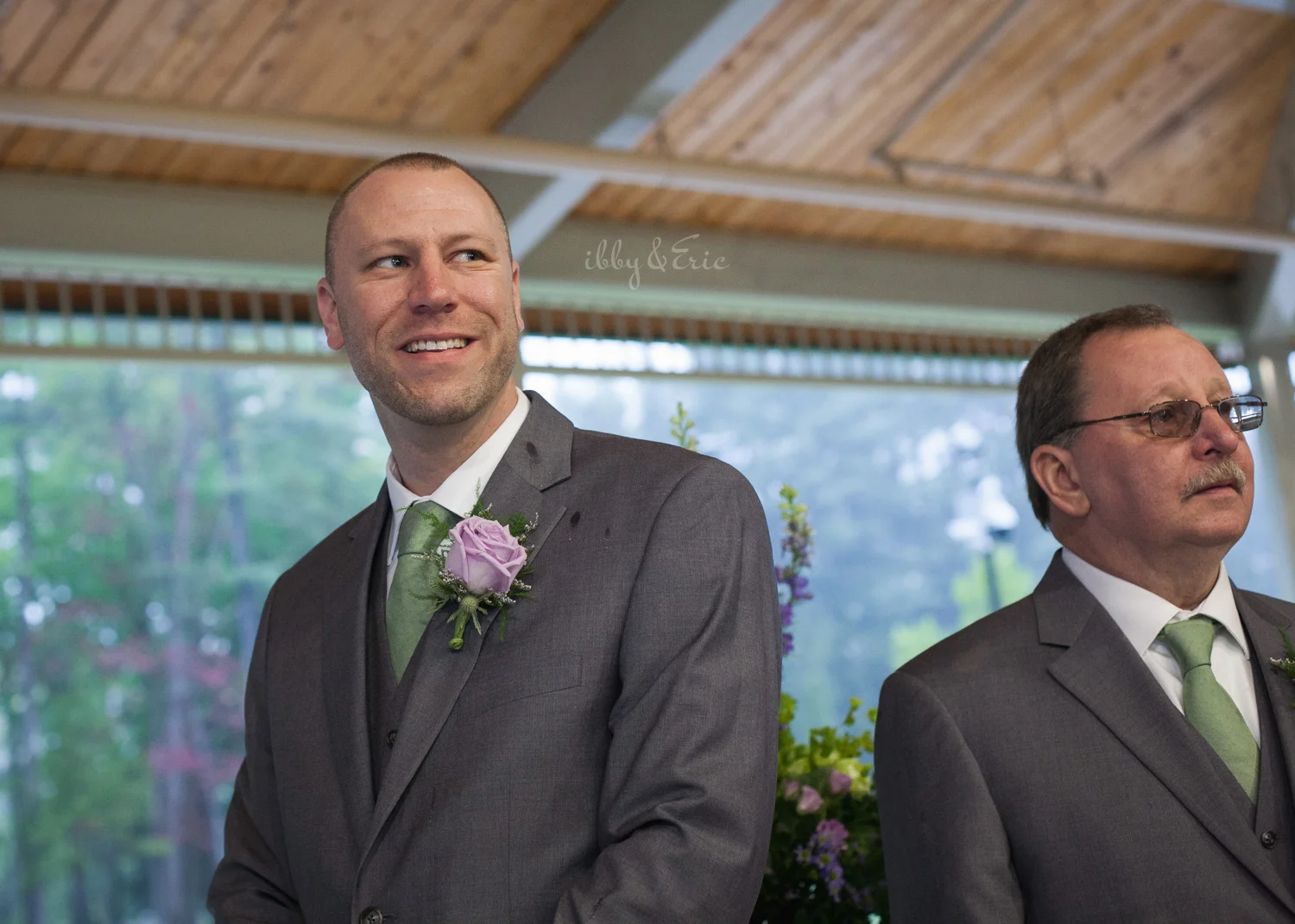 The groom wearing a gray suit and green tie smiles as he sees his bride walk down the aisle.