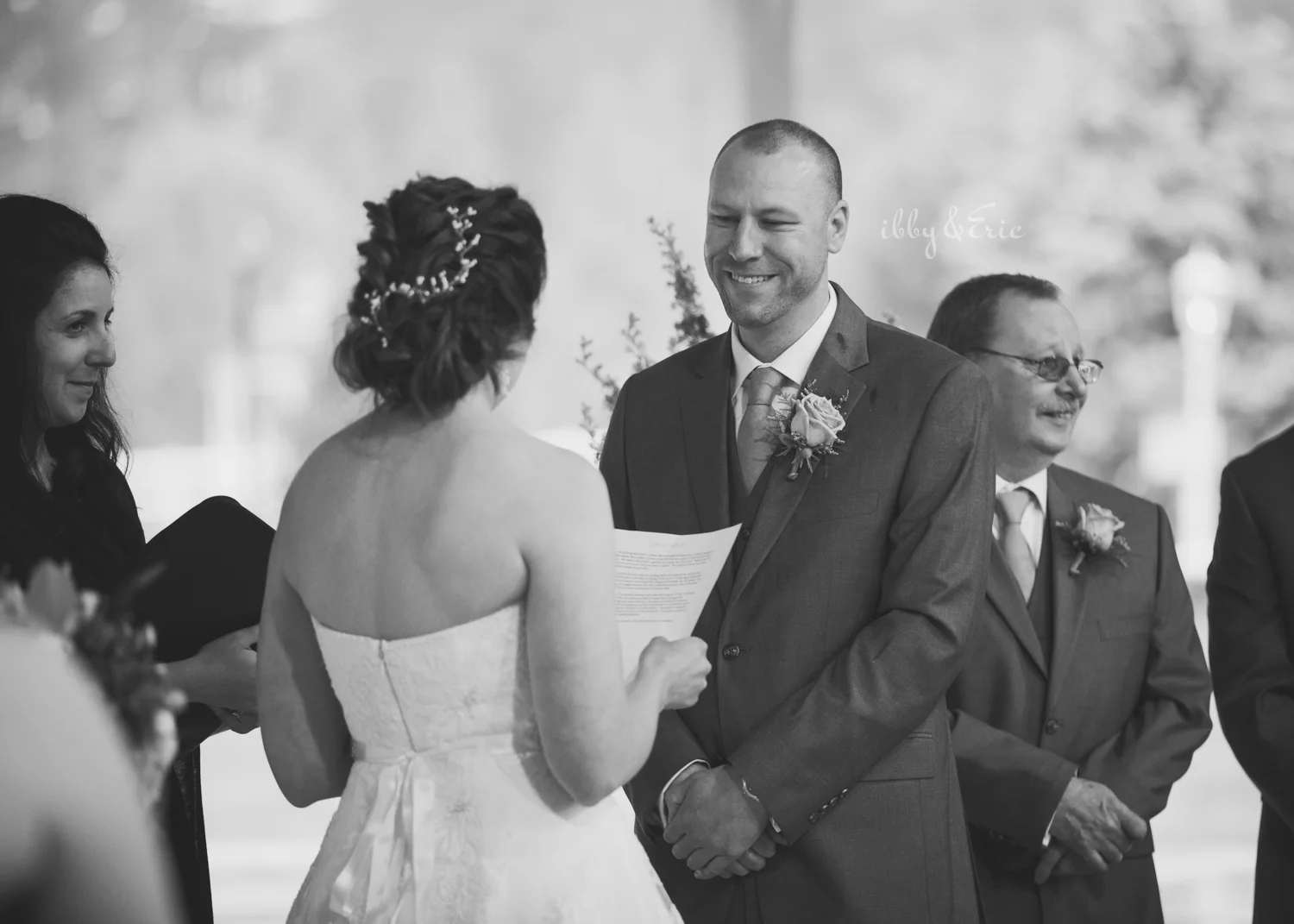 Black and white photo of the groom smiling as his wife reads her wedding vows.