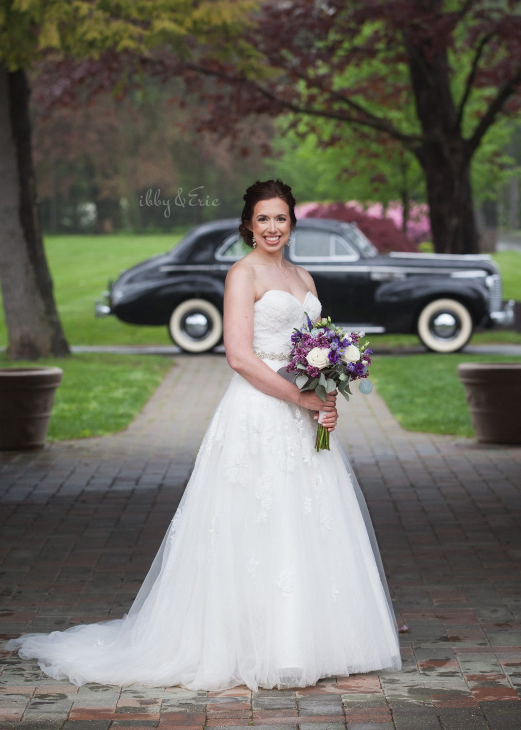 Smiling beautiful bride wearing a white lace wedding gown holds a purple bouquet in front of a vintage car.