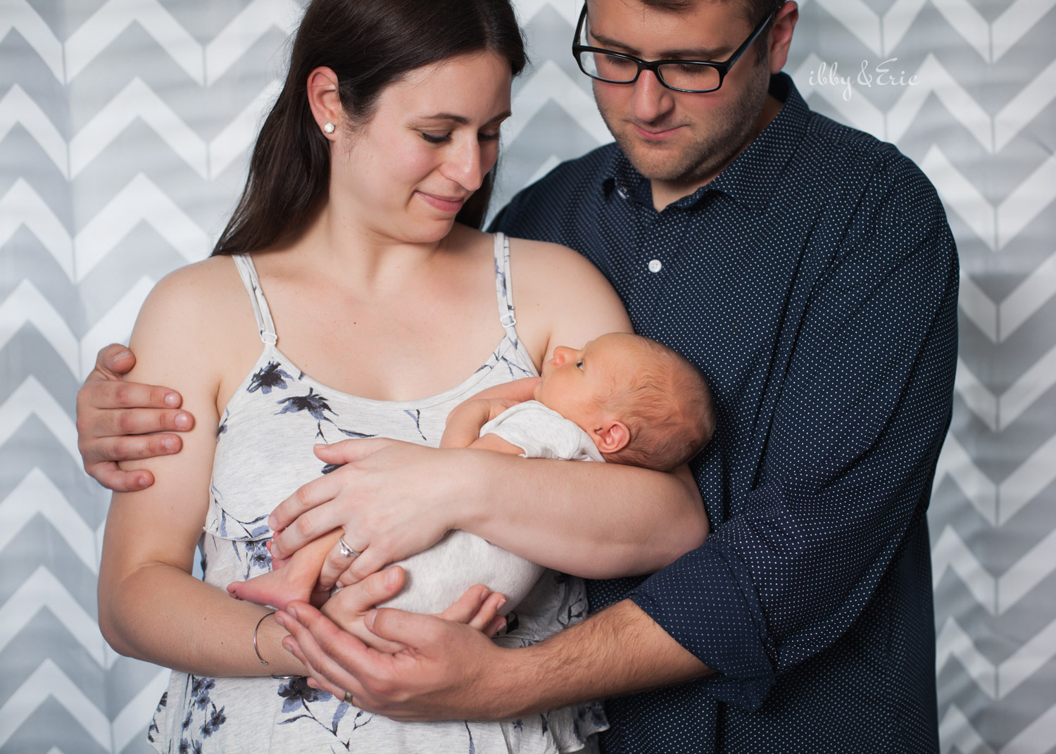 Mom and dad looking at their one week old baby boy in front of a gray curtain.