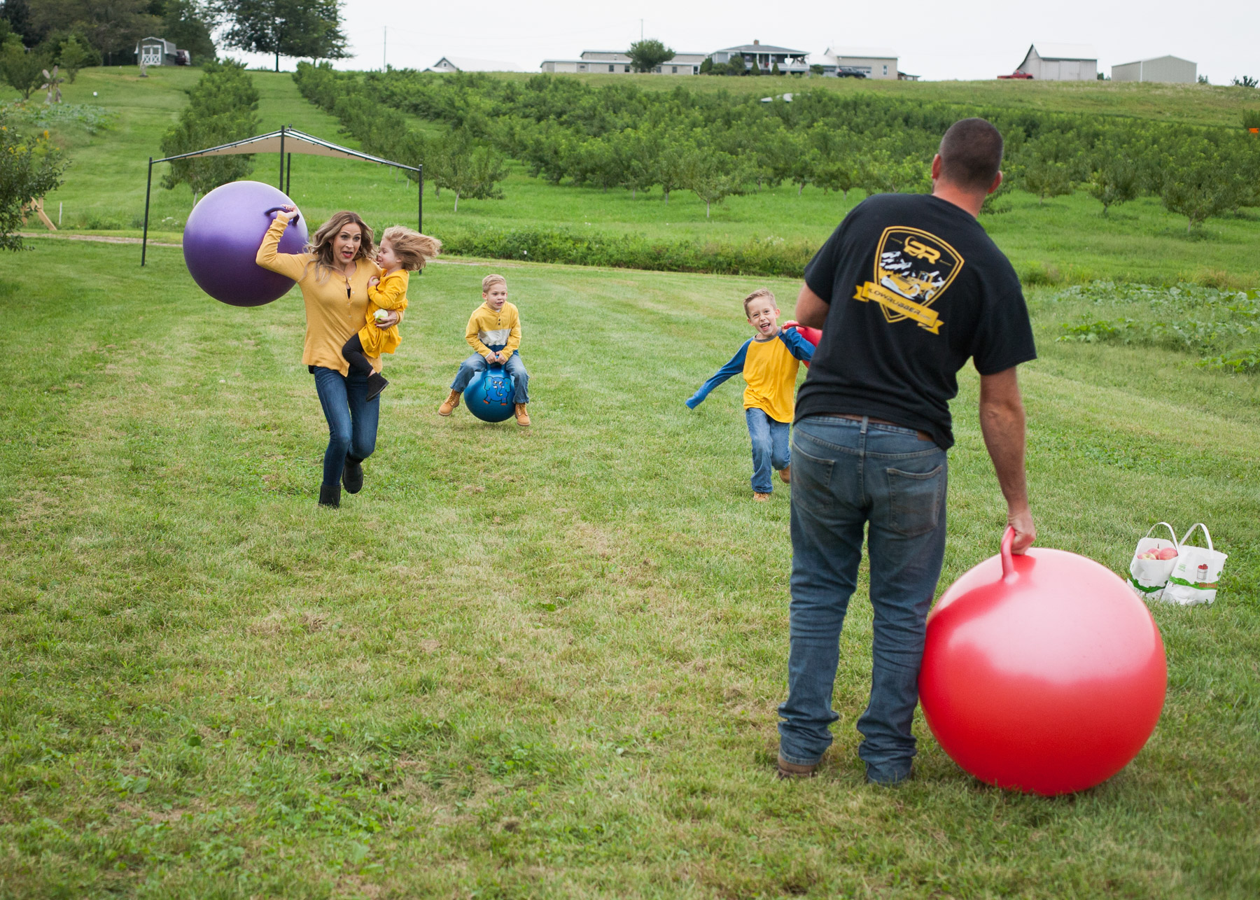 Mom holding her daughter in a yellow dress runs and throws a big bouncing ball at her husband.