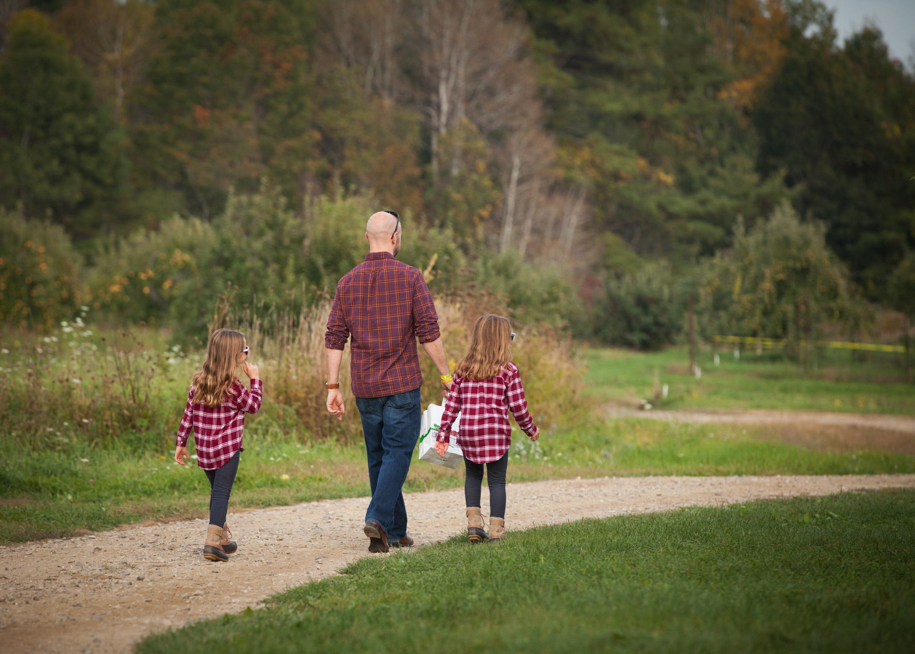 Dad and his daughters, all wearing jeans and red plaid flannel shirts, walk down a dirt path towards an apple orchard.