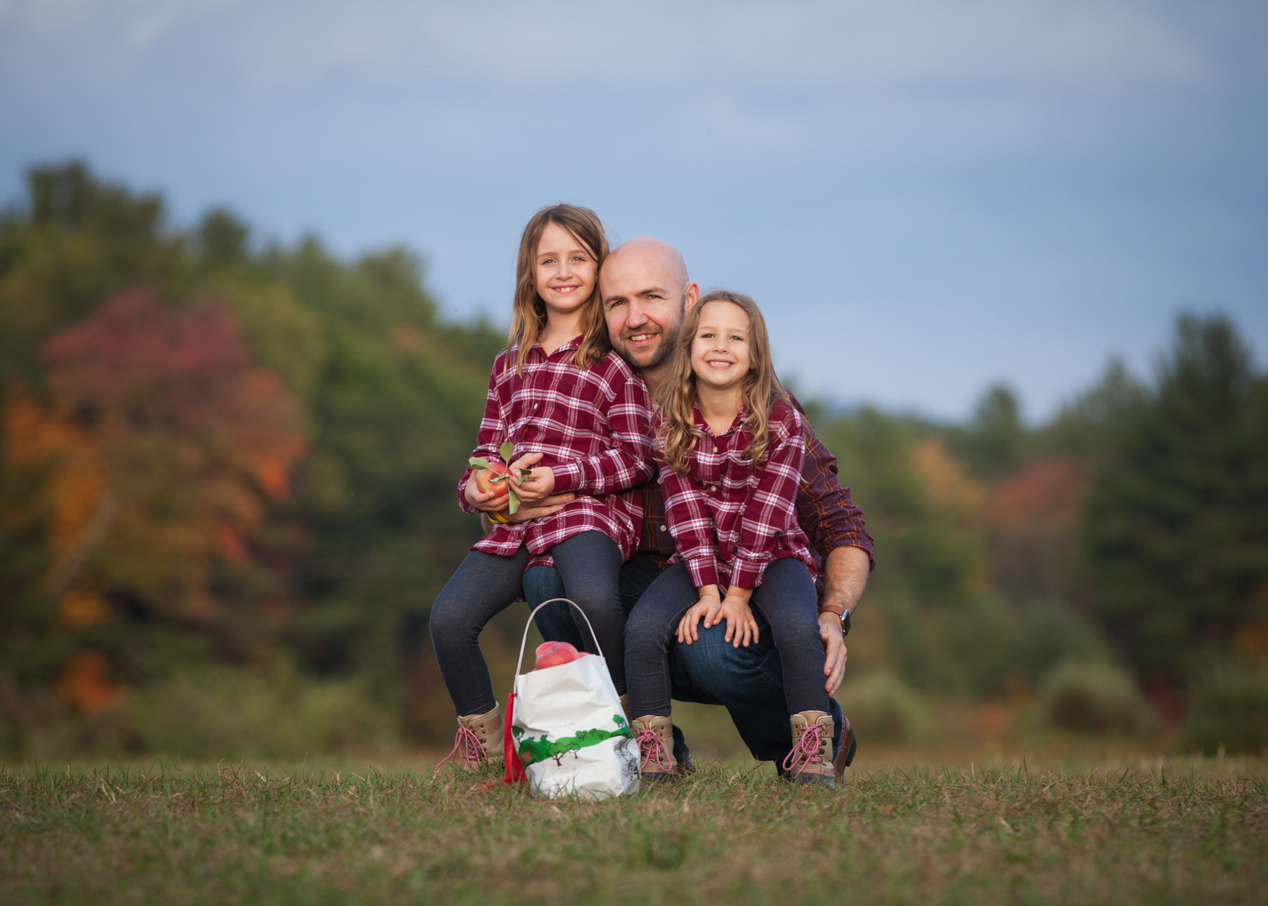 Dad and his daughters, wearing matching flannel shirts, sit together on a hill with a peck of apples in front and fall foliage behind them.
