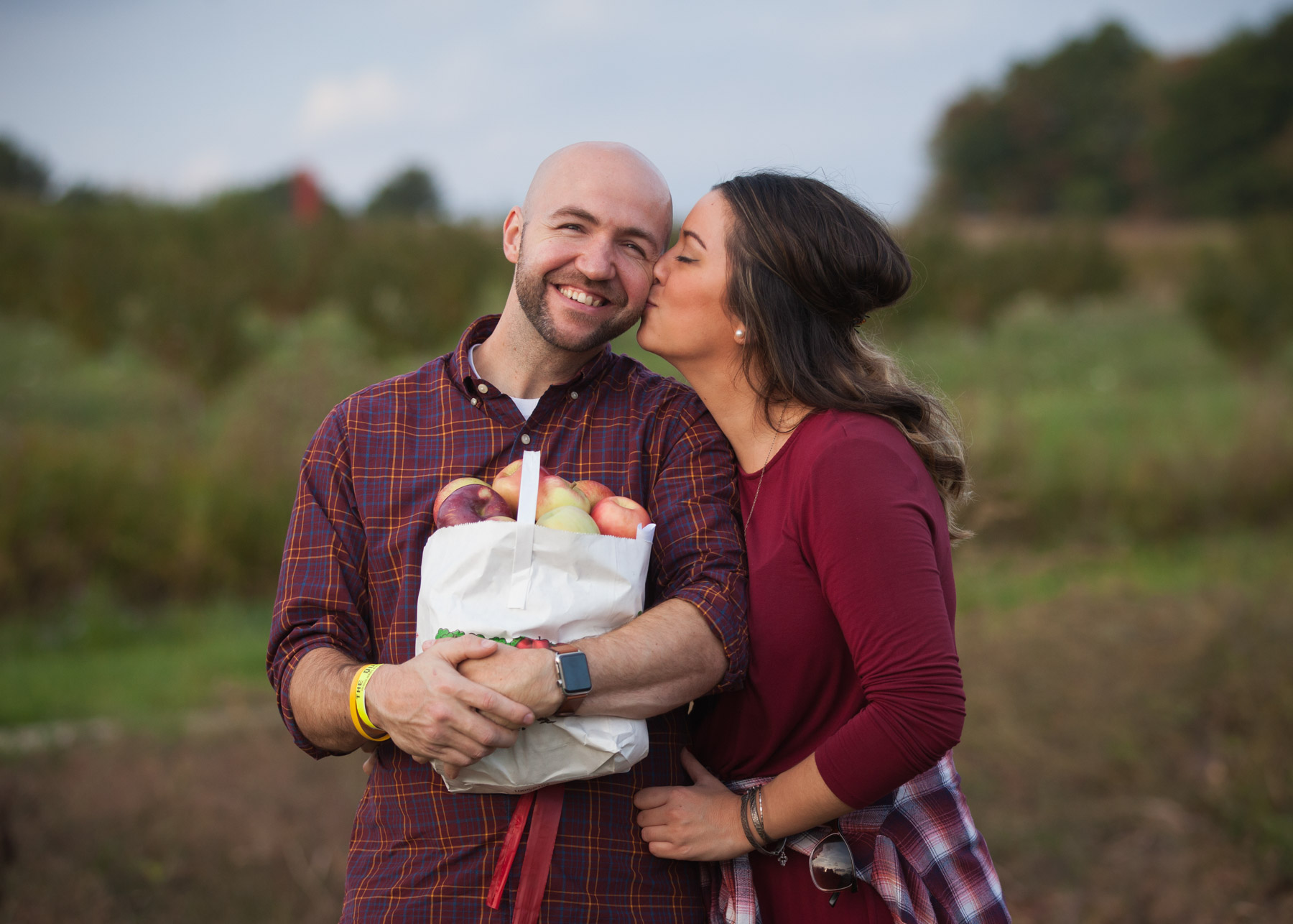 Woman with dark hair, wearing a burgundy shirt, kisses her boyfriend on the cheek while he smiles and holds a bag of apples.