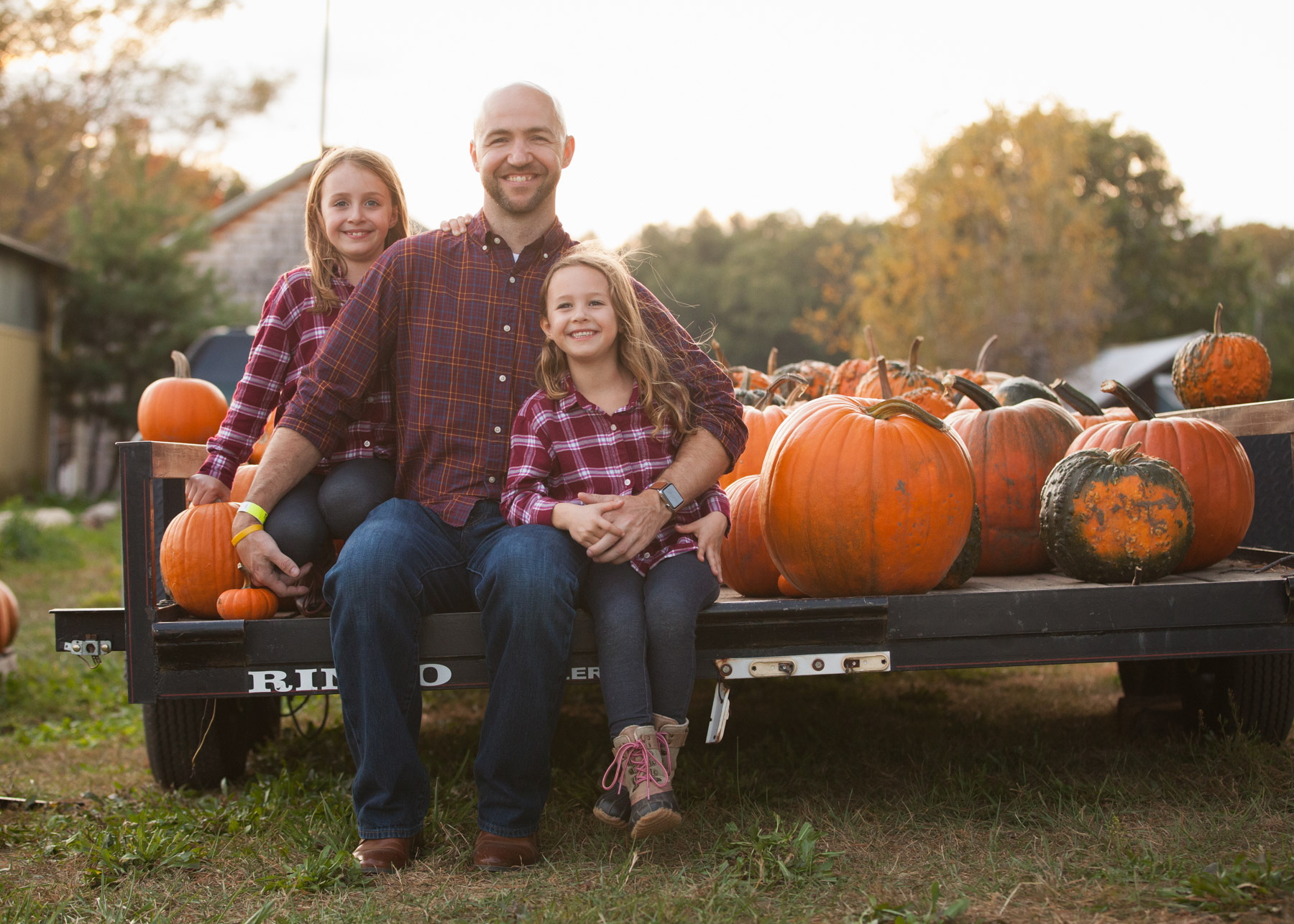 Dad and his two daughters sit on the back of a farm trailer filled with pumpkins just before sunset.
