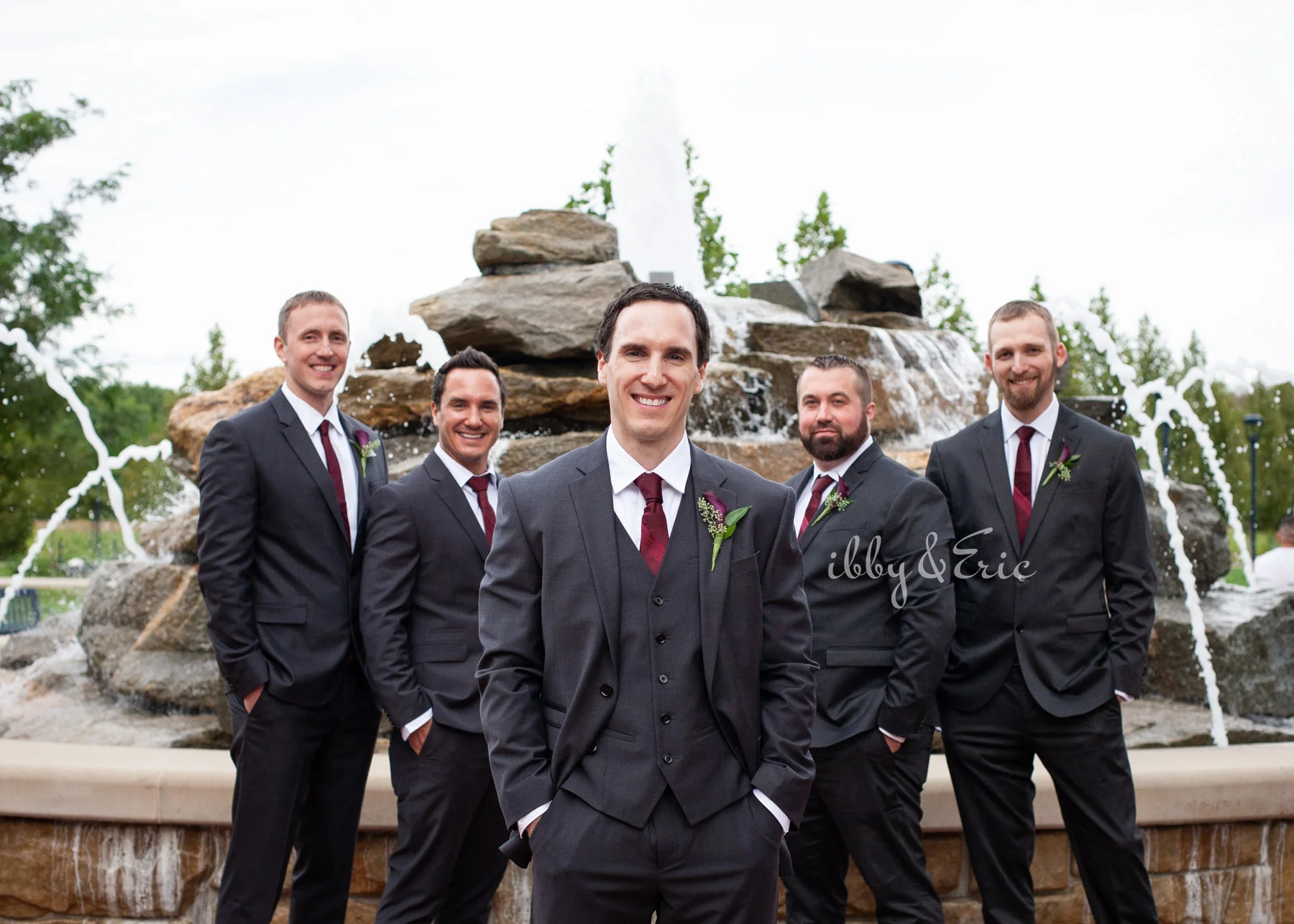 The groom stands in front of a fountain in Glastonbury with his 4 groomsmen wearing black suits with burgundy ties.