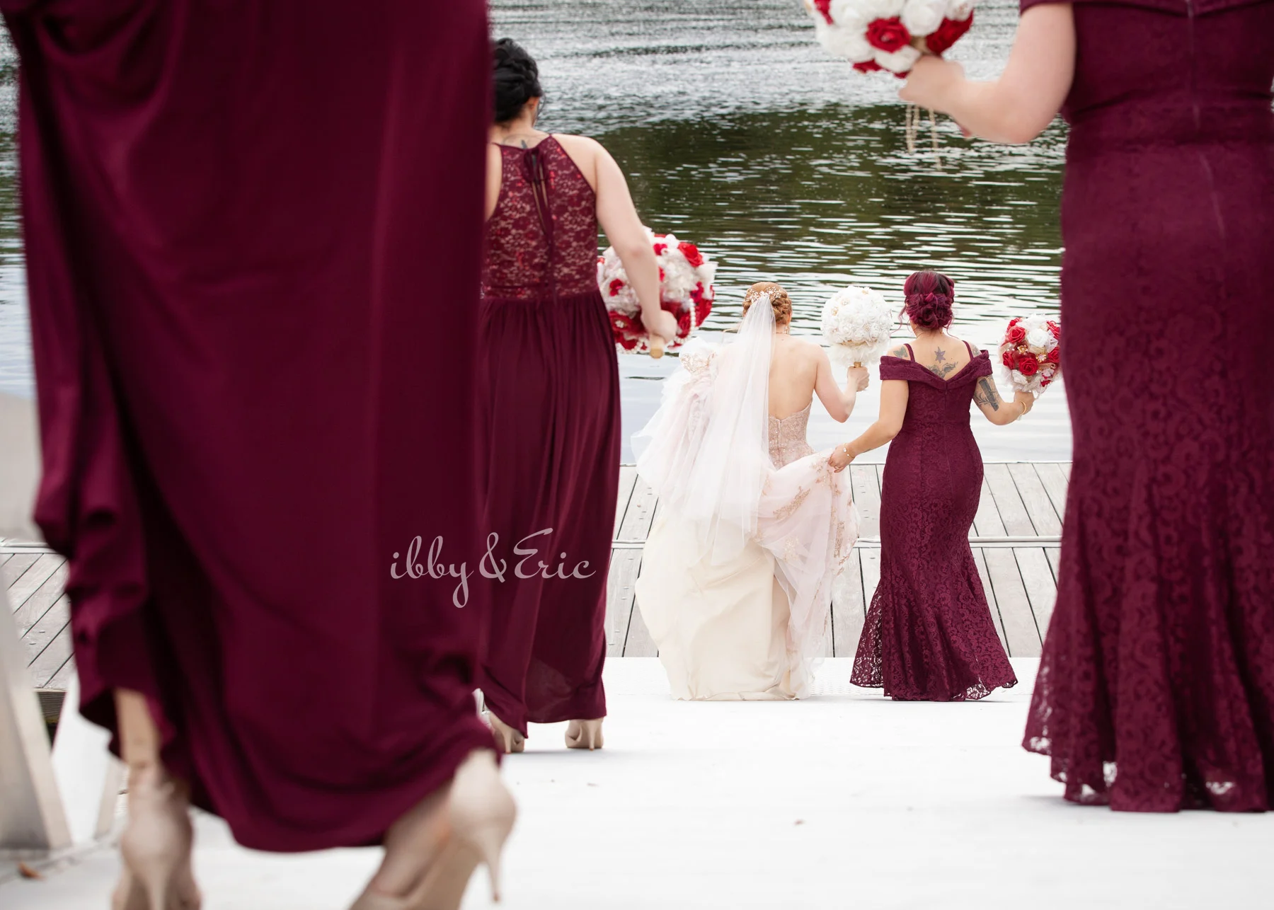 Bride walks down a riverfront dock with her bridesmaids wearing burgundy lace dresses.