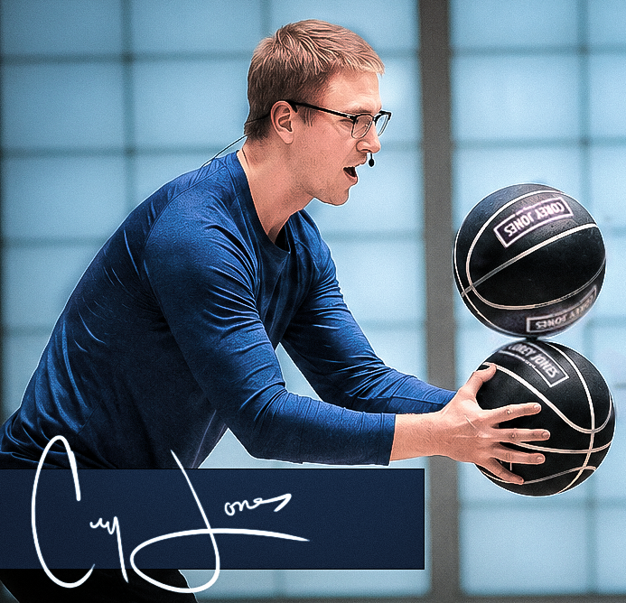 A man in a blue shirt and glasses is playing basketball indoors with two basketballs in mid-air.