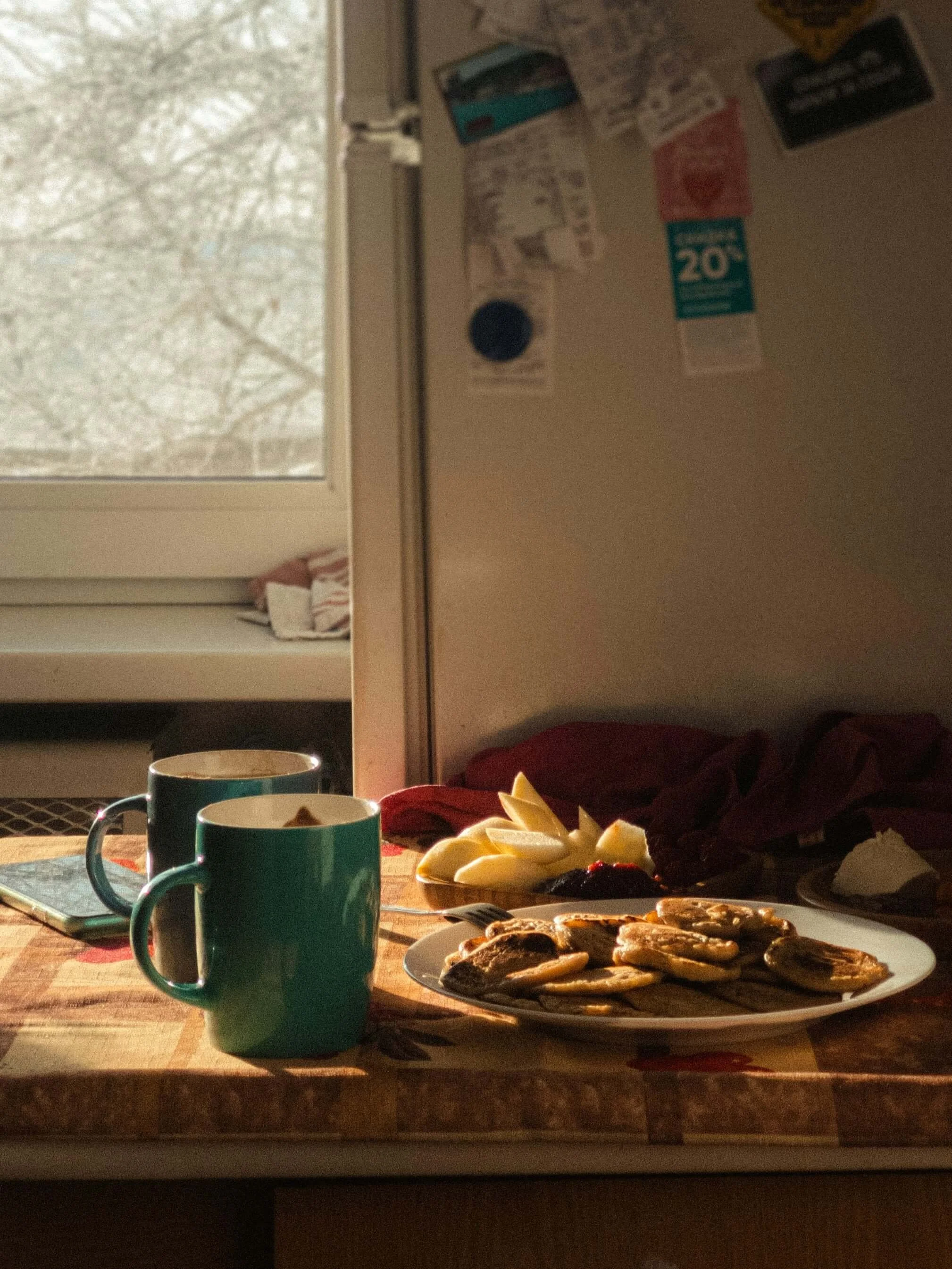 Breakfast spread with pancakes, fruit, and two mugs on a wooden table near a window.