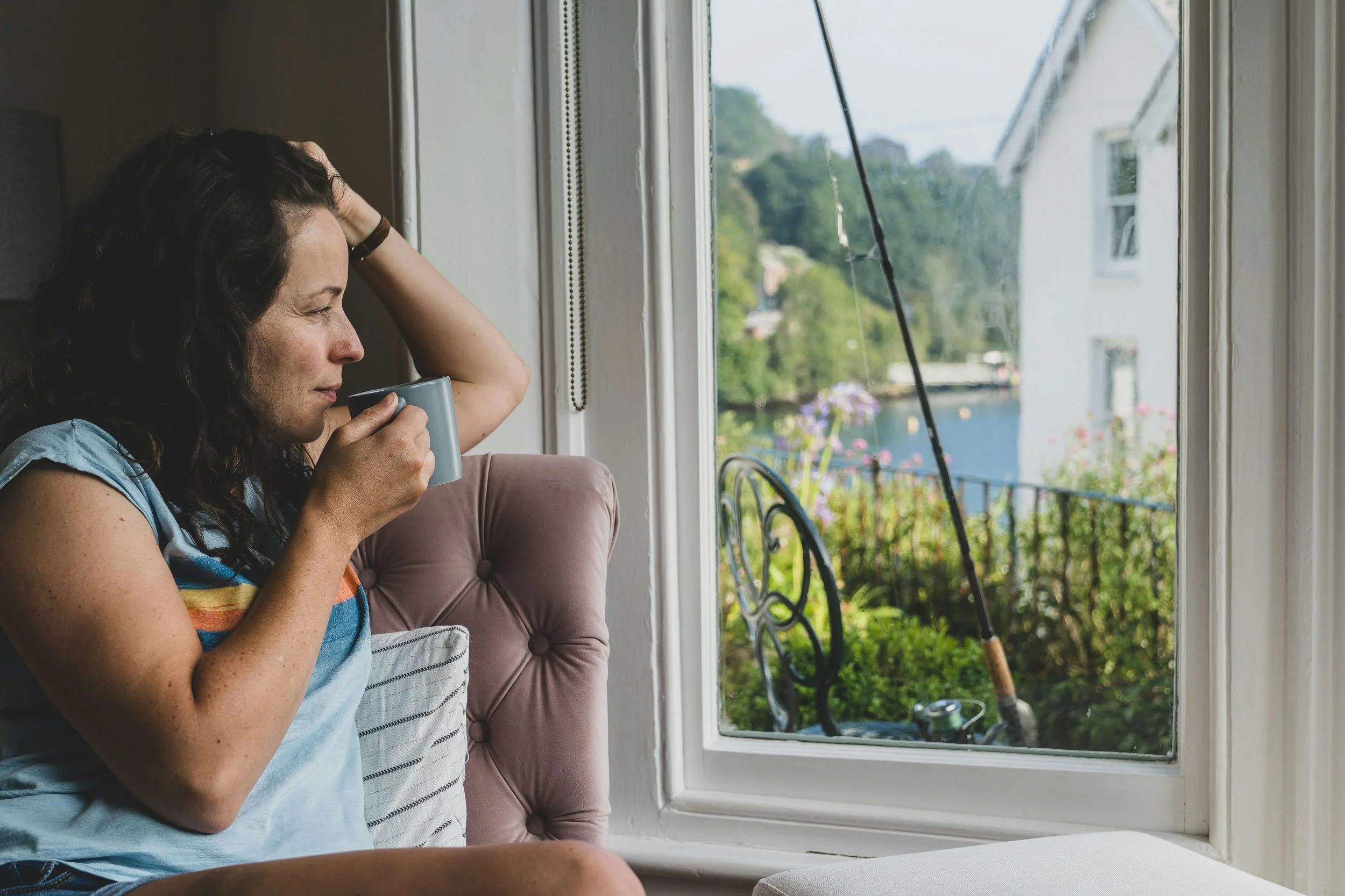 Woman sitting by a window with a mug, reflecting on the discomfort of feeling full after eating.
