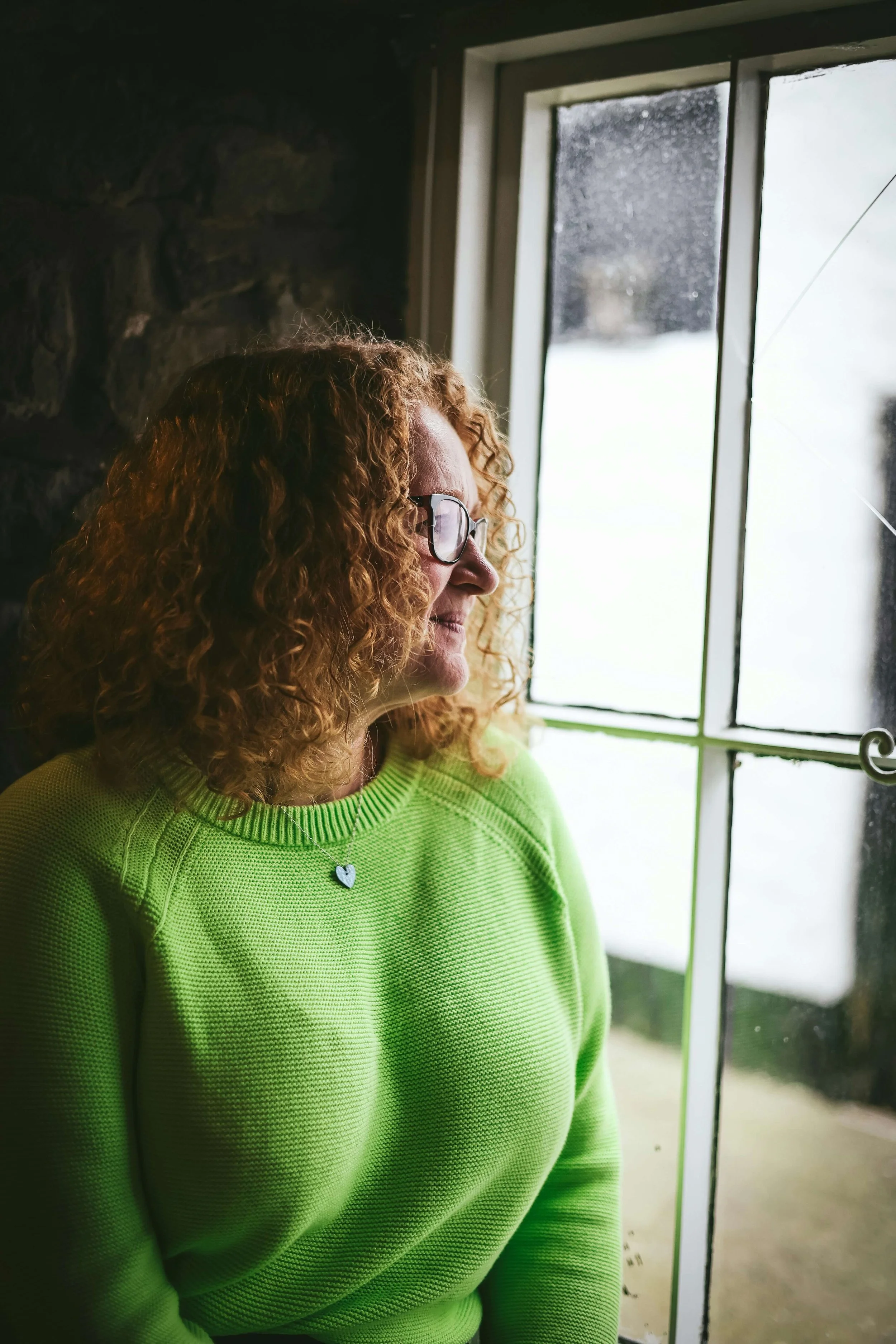 Woman in a green sweater looking out a window in natural light.