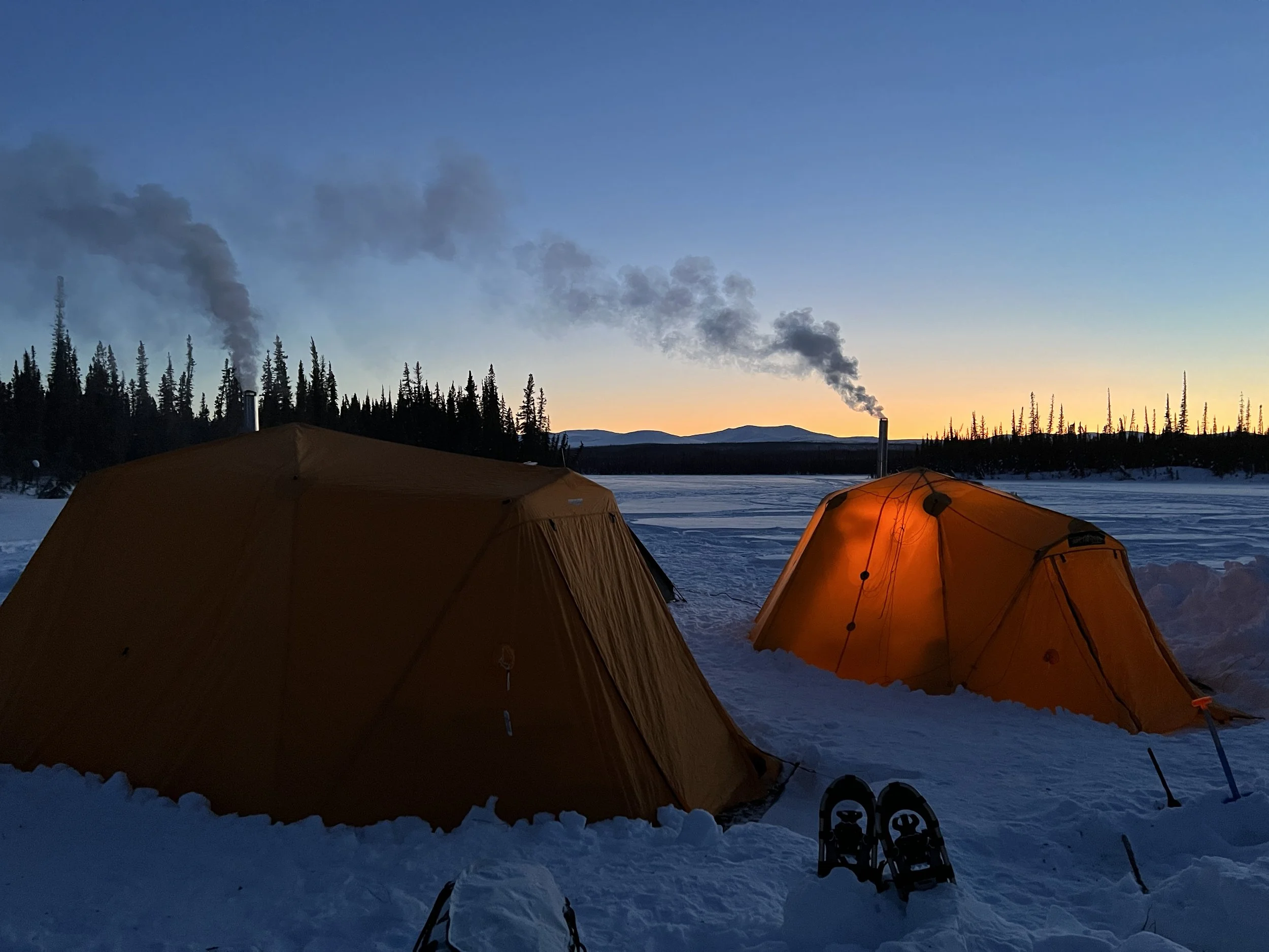 Two orange tents set up in a snowy landscape during sunset with smoke rising from chimney stacks and snowshoes placed nearby.