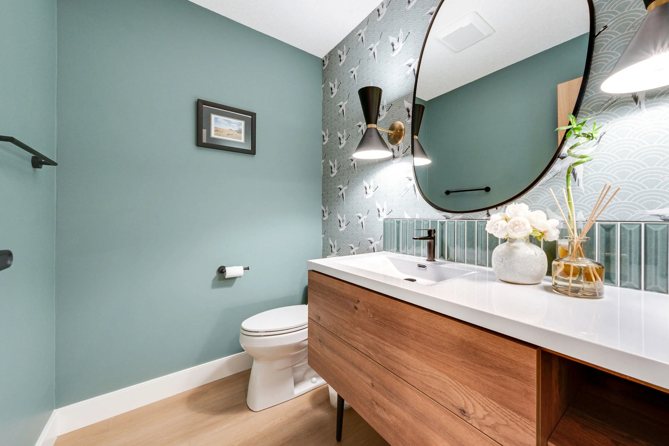 A modern bathroom with a light turquoise wall, a wooden vanity with a white countertop, a round mirror, and decorative vases with flowers and reeds. There is wall art and black sconces with white lights.