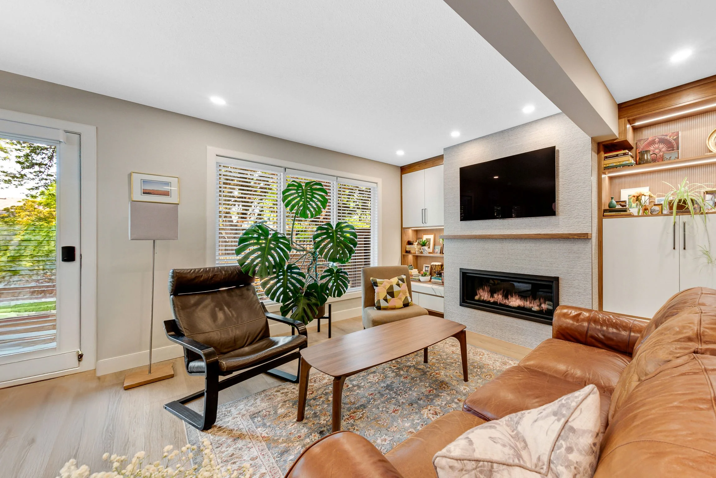 Living room with leather sofa, armchair, coffee table, large houseplant, fireplace, wall-mounted TV, shelves with decor, and windows with blinds