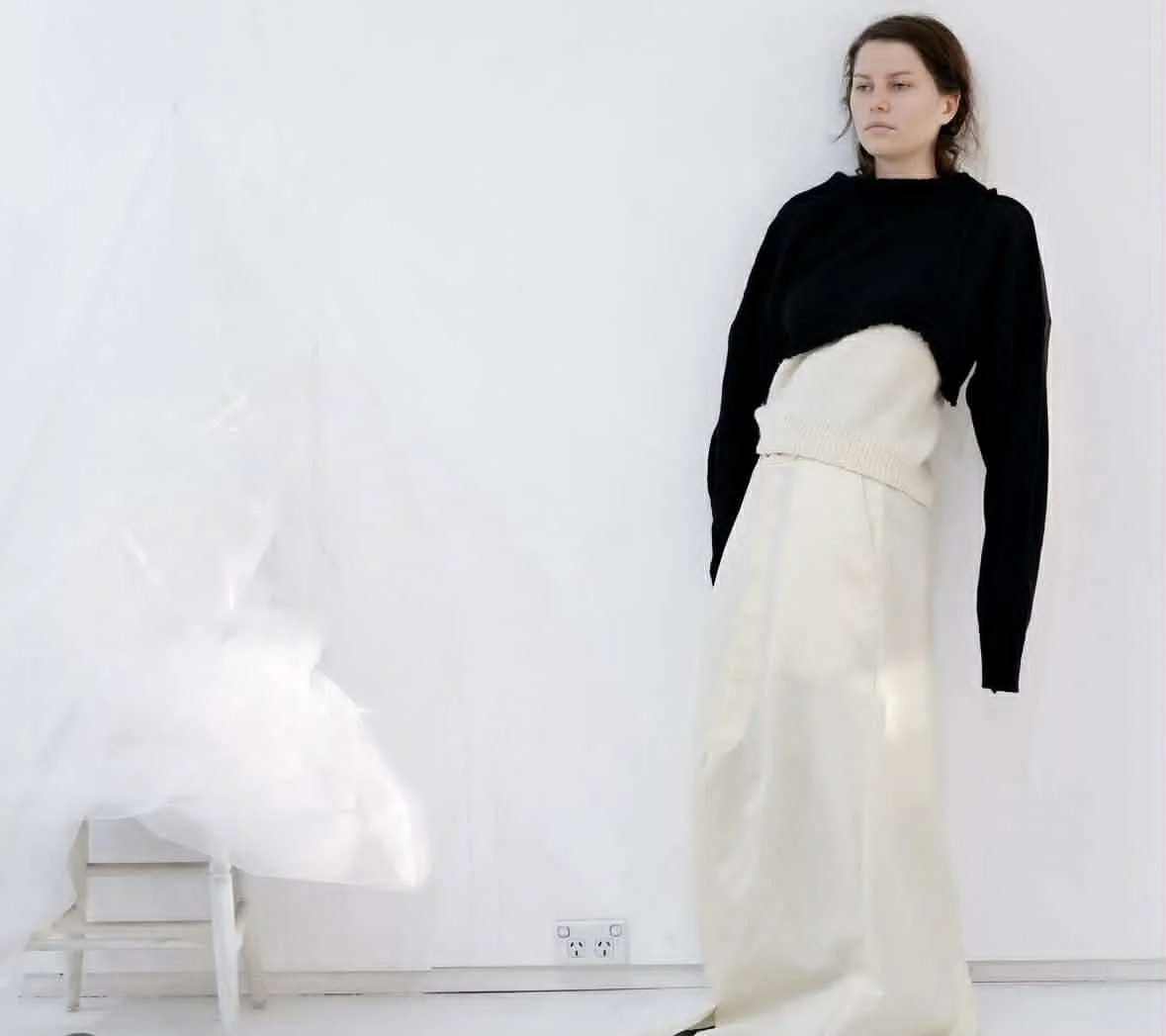 A woman standing in front of a white wall, wearing a black long-sleeve top and cream-colored high-waisted wide-leg pants.