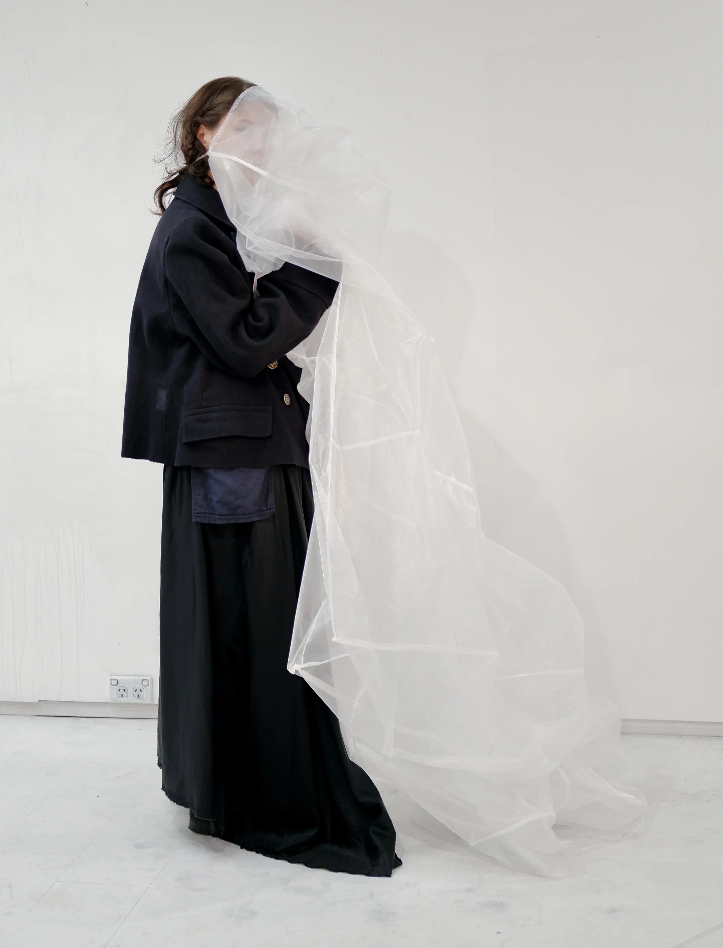Person with brown hair wearing a black jacket and dress, holding a transparent plastic sheet against a plain white wall.