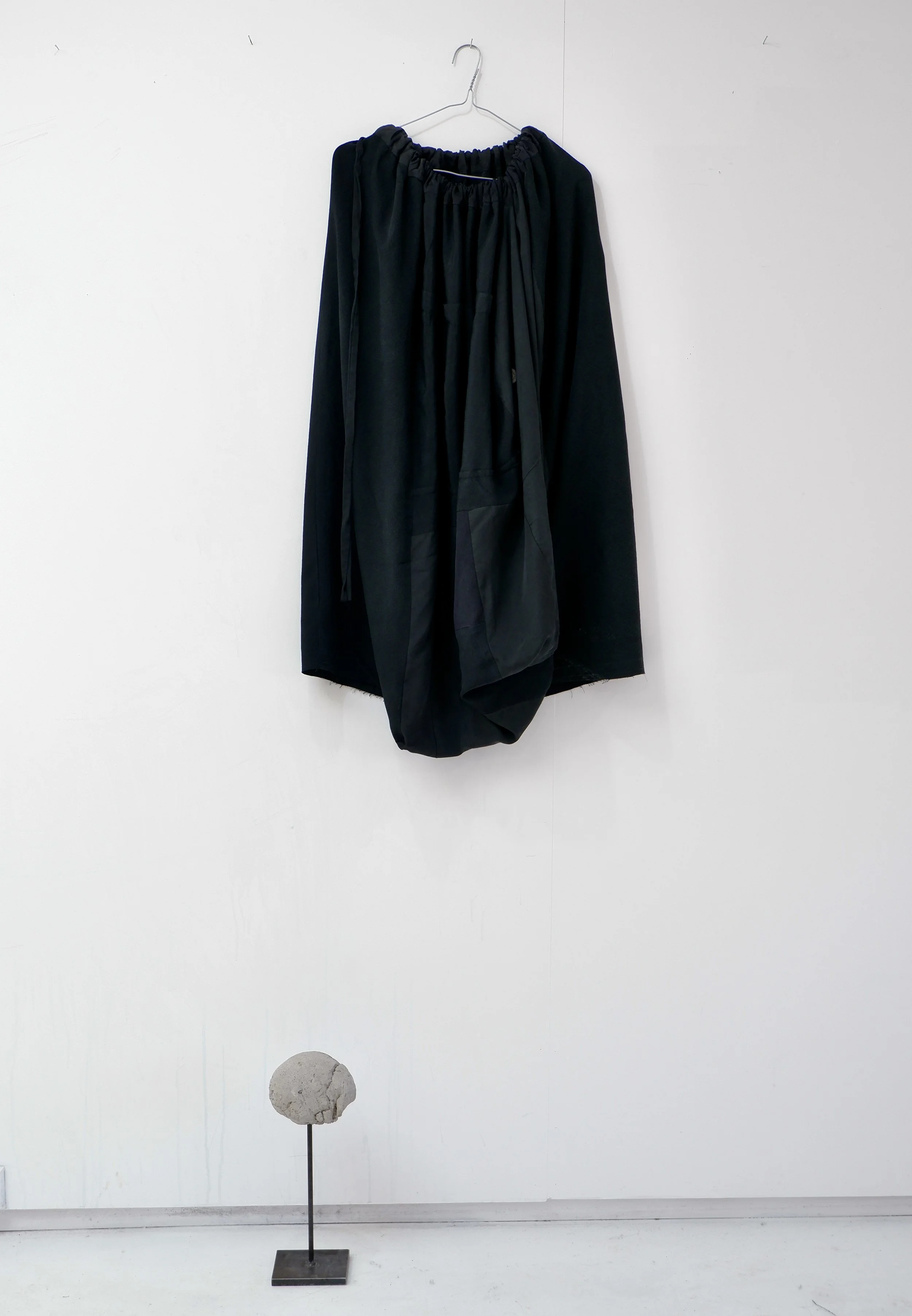 Black clothing garment hanging on a wire hanger on a white wall, with a small sculpture of a rock on a stand on the floor below.