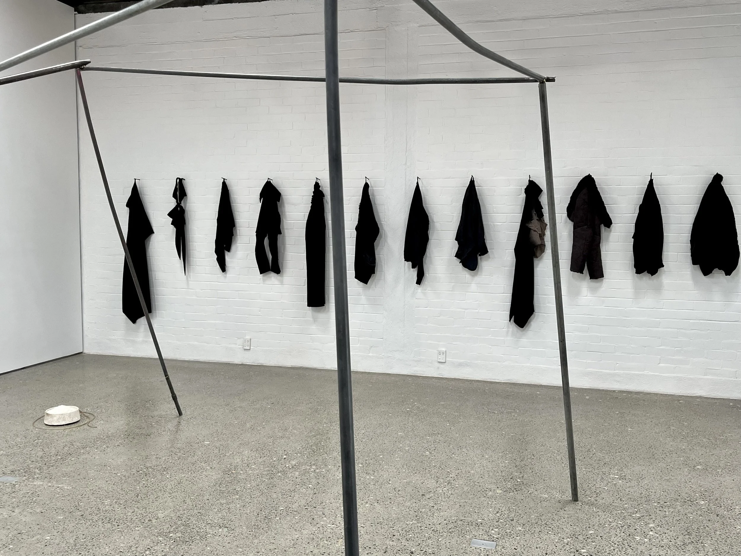 A white brick wall with black clothing items hung on hooks and a sculpture-like metal structure in the foreground.
