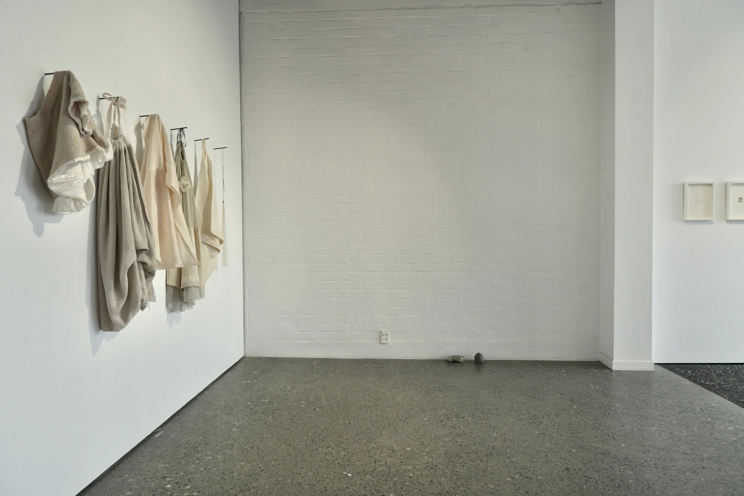 An art gallery with beige and cream-colored clothing and accessories hanging on the left wall. The center has a white brick wall and a gray speckled floor. There are a few small rocks or objects near the wall socket at the bottom.