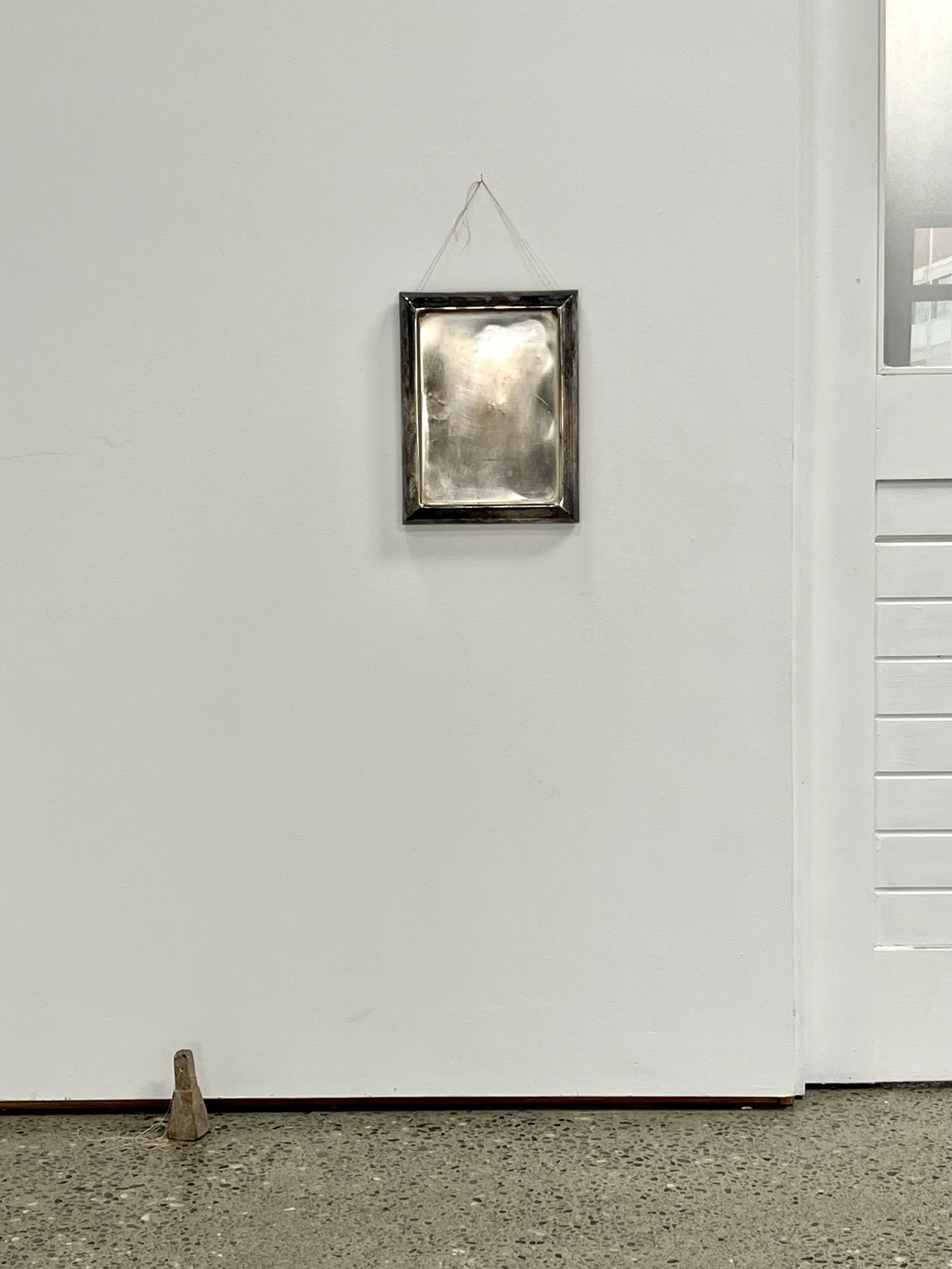 Photo of a plain white wall with a small empty silver picture frame hanging from a string in the center and a small cone-shaped object on the floor near the left bottom corner.