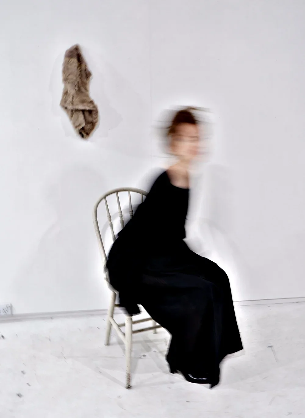 Blurry woman in black sitting on a white wooden chair in front of a white wall with a hole and brown fur or fabric on it.