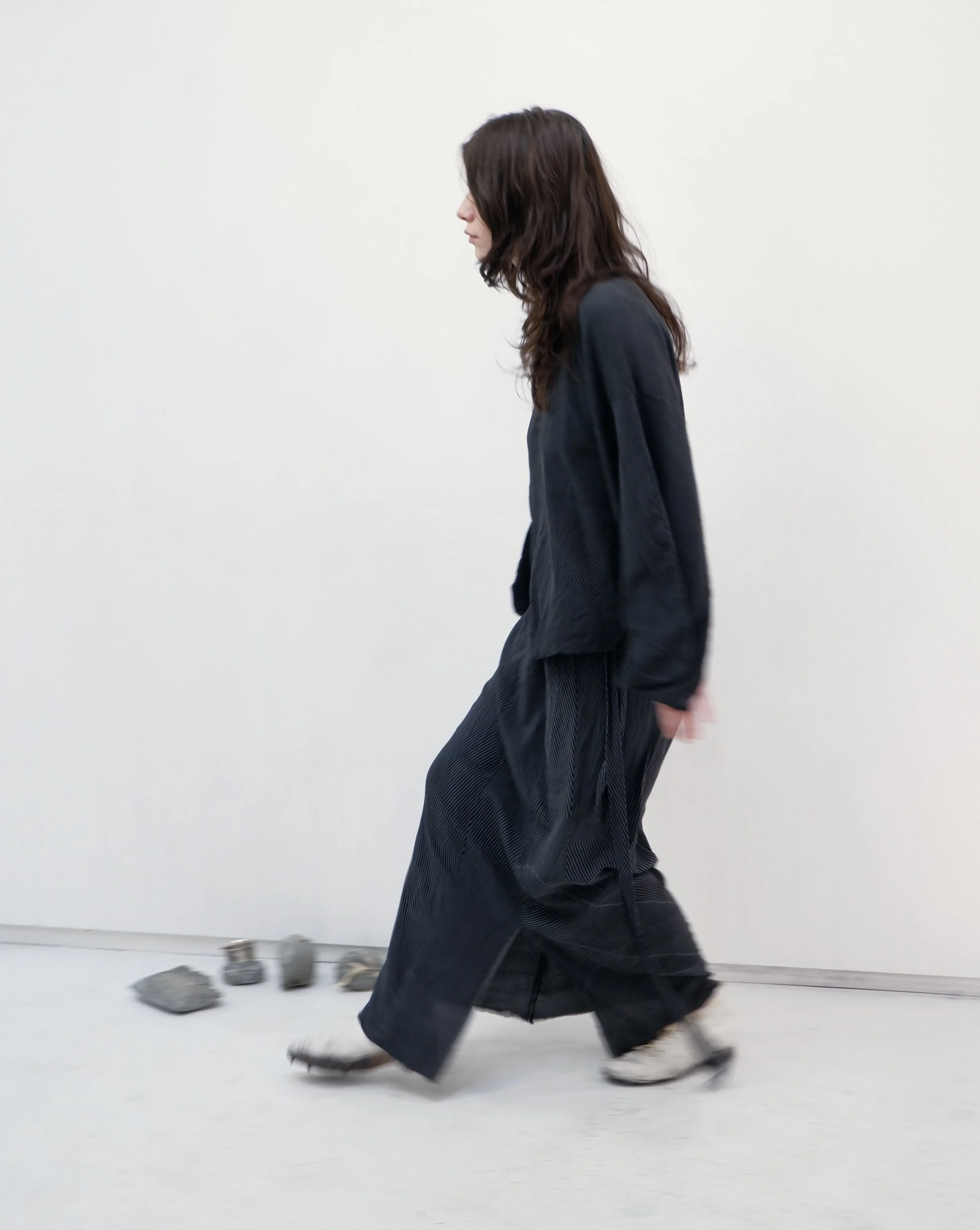 A woman with long brown hair walking on a white floor with rocks and small objects behind her, wearing a black outfit and skates.