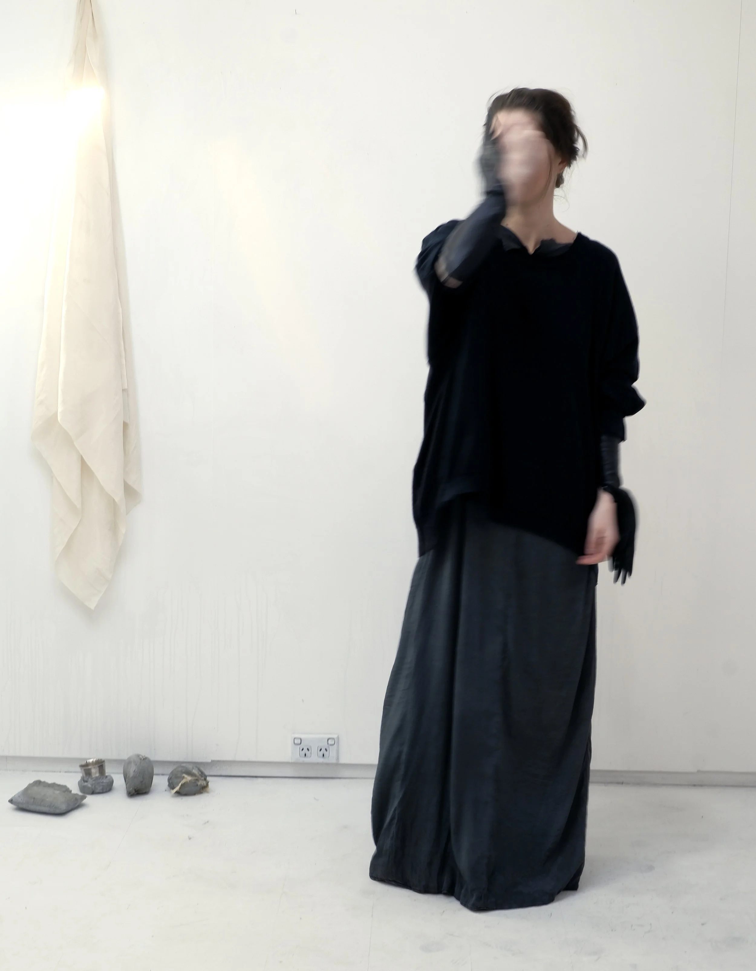 A woman standing indoors against a blank white wall, wearing a long black skirt, black top, and black gloves, covering her face with her right hand while her left hand rests by her side.