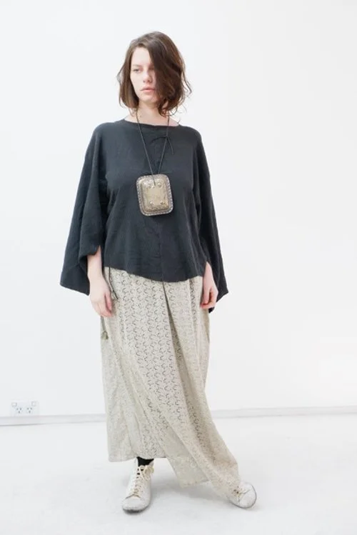 A woman with shoulder-length brown hair wearing a loose black top, patterned beige wide-leg pants, white sneakers, and a small silver purse on a long chain.