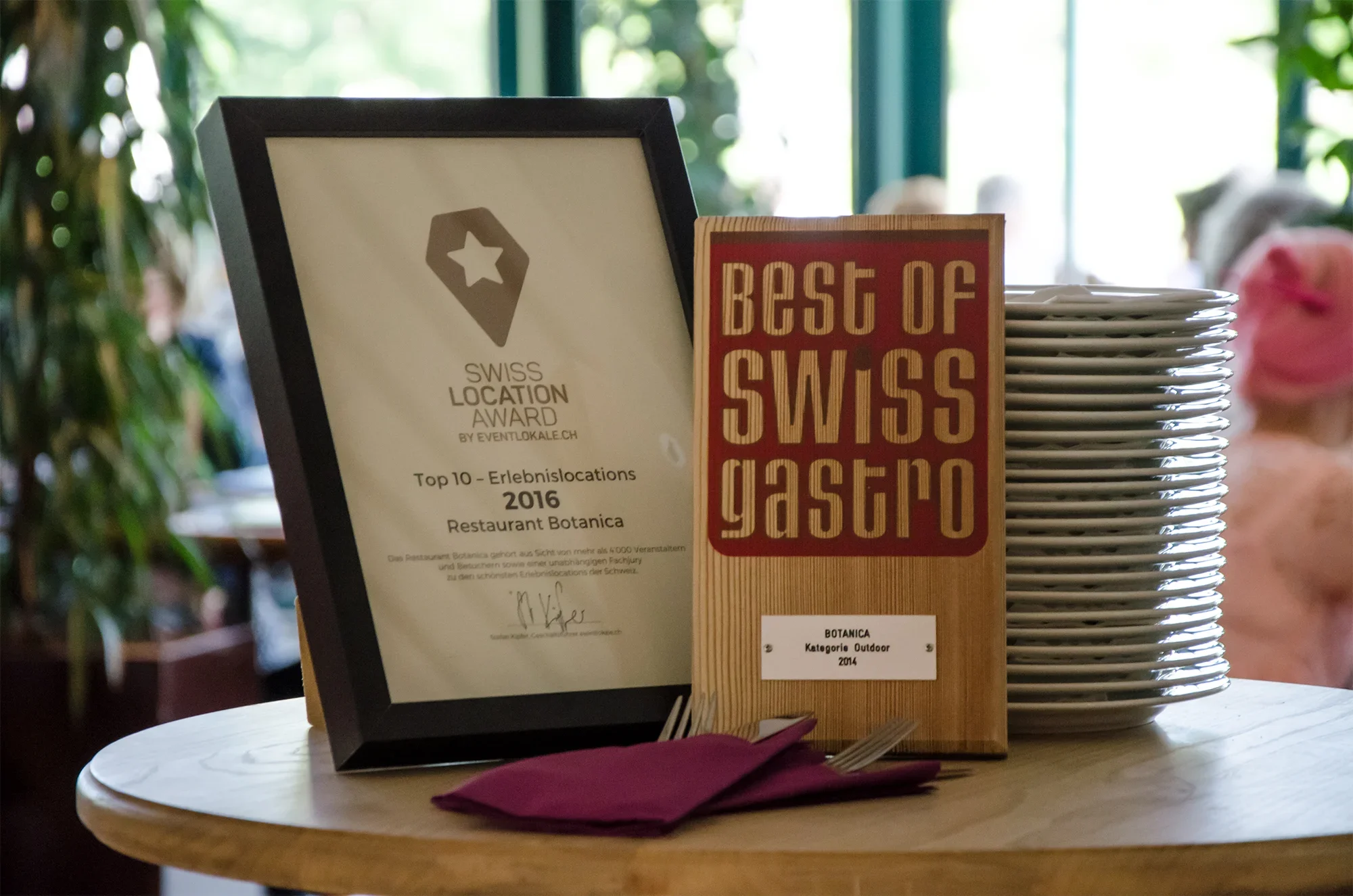 Certificate of recognition, stack of white plates, and a sign with the text 'Best of Swiss Gasto' on a wooden stand on a round table inside a restaurant with green plants in the background.