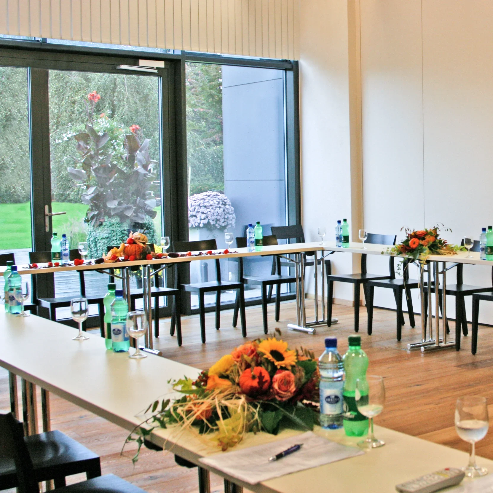 A room set up for a meeting or event with tables, chairs, floral arrangements, water bottles, and glasses, with large windows showing greenery outside.