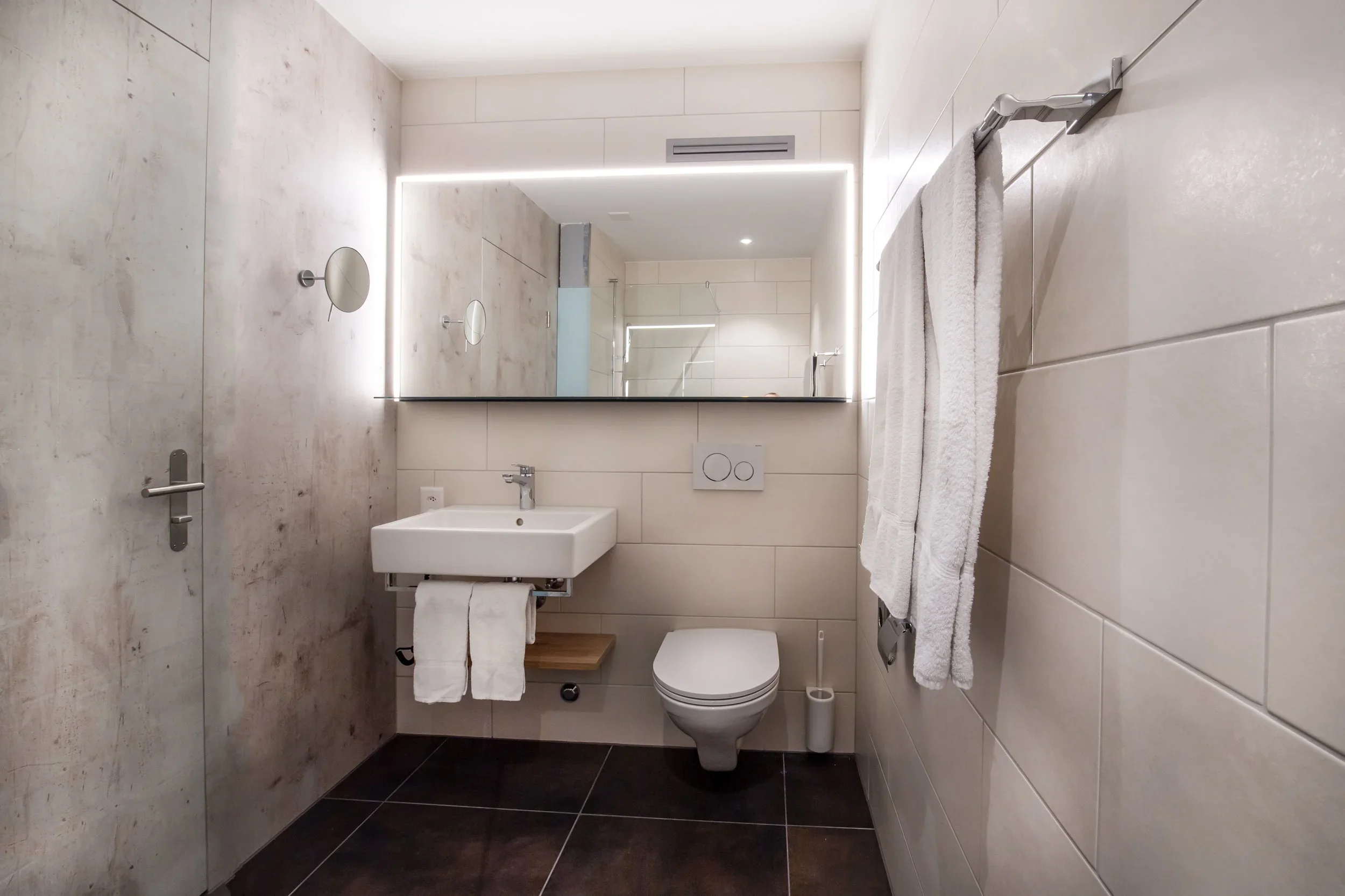 Modern minimalist bathroom with beige tiled walls and black tiled floor. Features a wall-mounted toilet, a white sink with a towel rack, and a large mirror with LED lighting. Towel bars and small mirror on the wall.