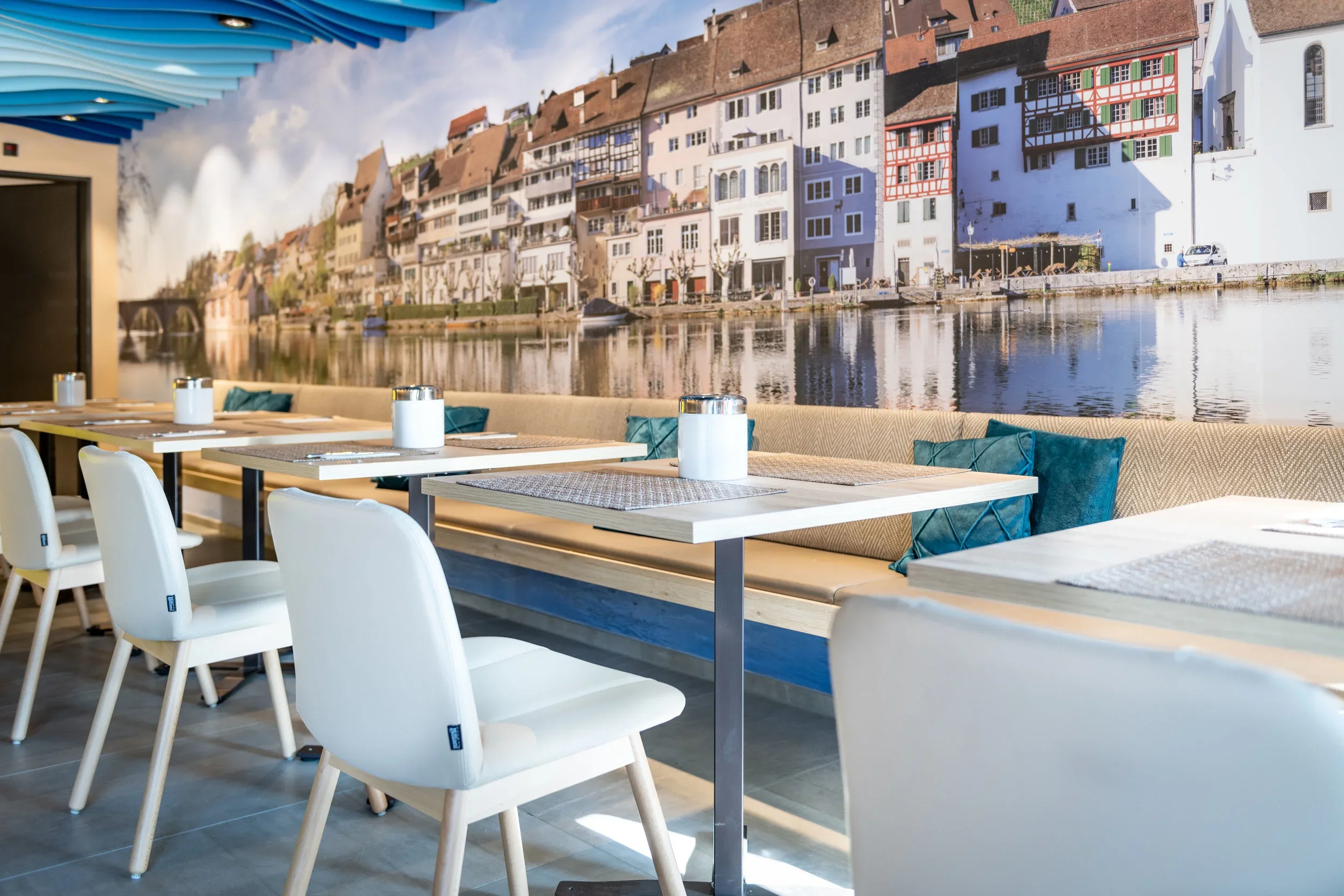 Interior of a restaurant with a large mural of a European canal scene on the wall, showing colorful historic buildings along the water, with benches and tables set for dining.