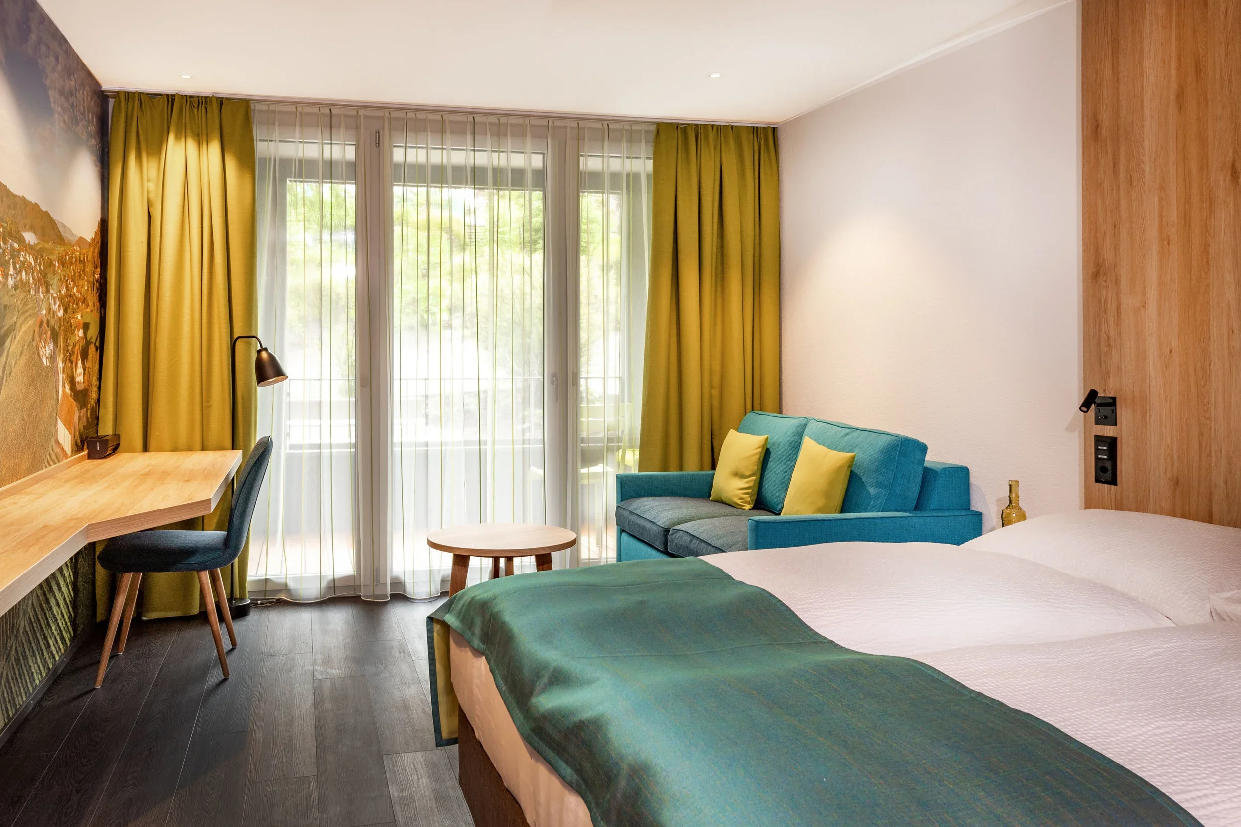 A hotel room with two beds, a turquoise sofa, a wooden desk with a black chair, yellow curtains, and large windows letting in natural light.