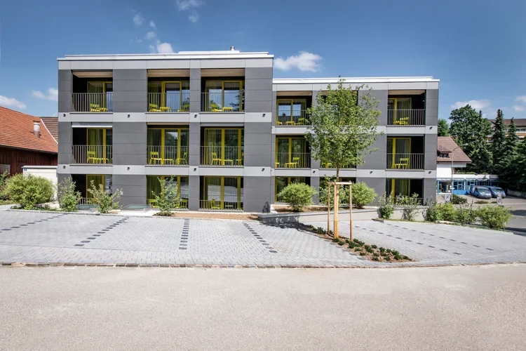 Modern three-story apartment building with gray exterior, yellow railings, and small balconies. Surrounding landscaping includes young trees and bushes. In front, there is a parking lot with designated spaces and a paved road.