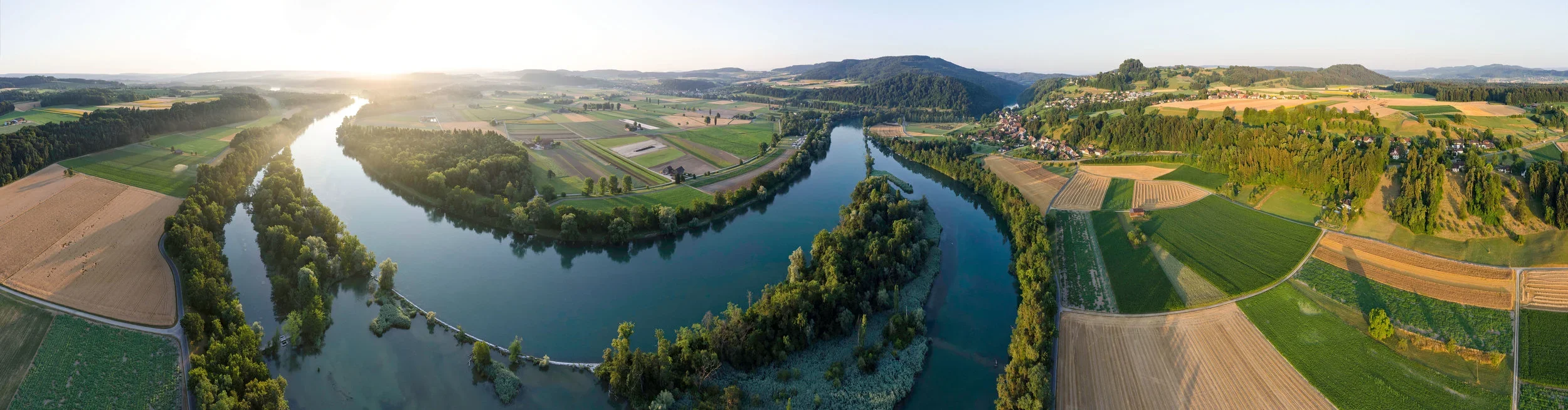 Aerial view of a winding river surrounded by green forests and farmland, with hills in the distance at sunset.