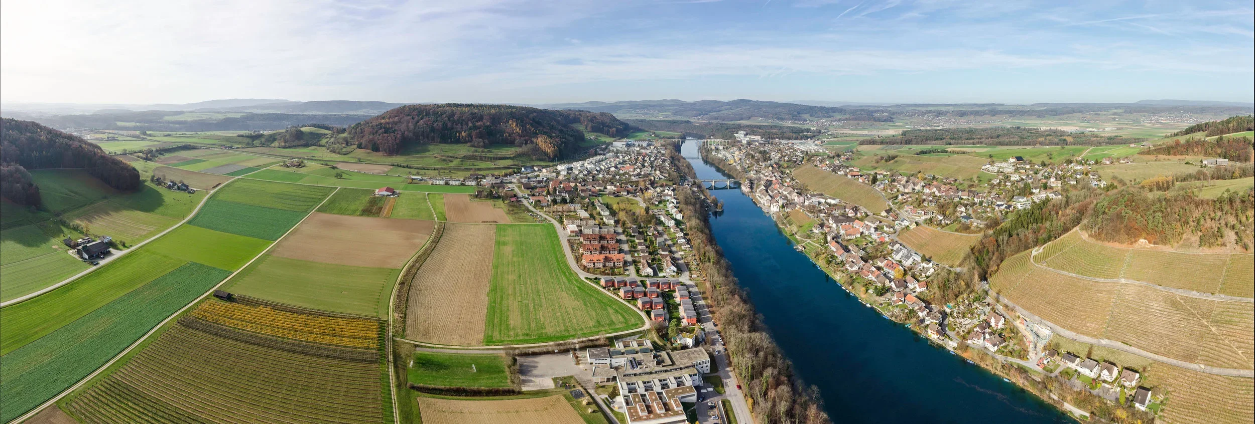 Aerial view of a town situated along a winding river, with surrounding farmland and hills under a partly cloudy sky.