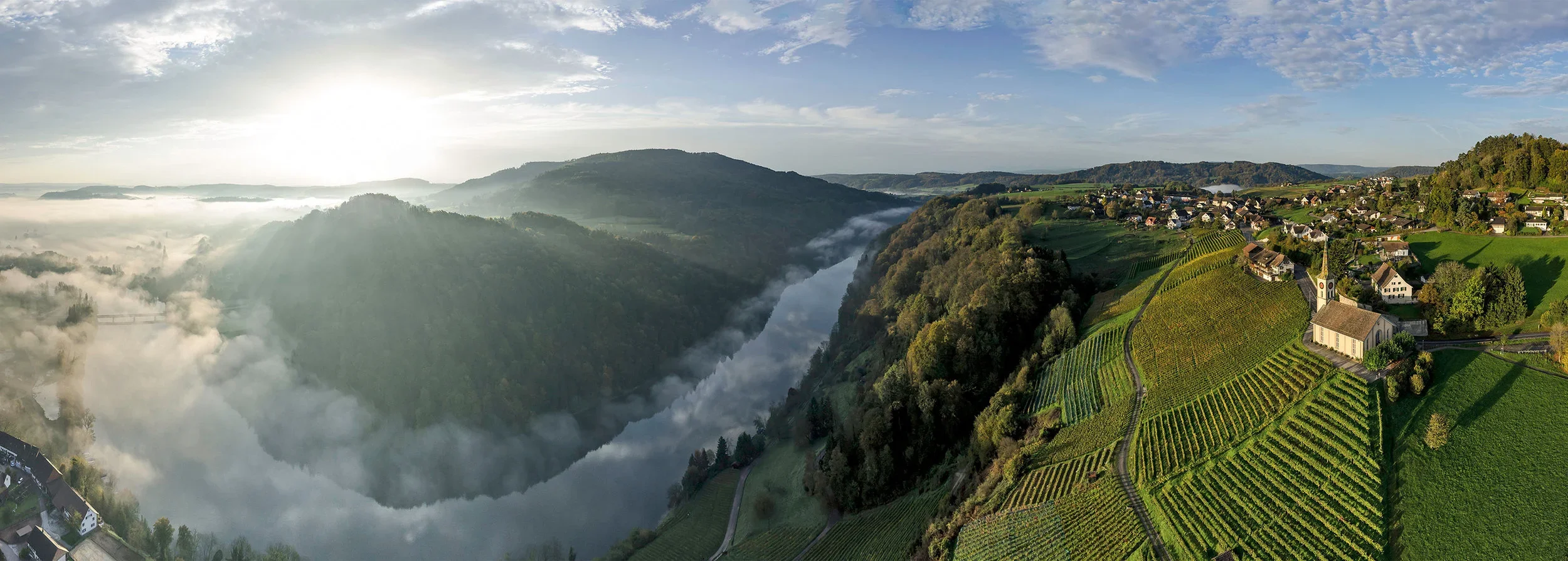 An aerial view of a river running through a lush green valley with surrounding wooded mountains and a small village with vineyards on the hillside, under a partly cloudy sky.
