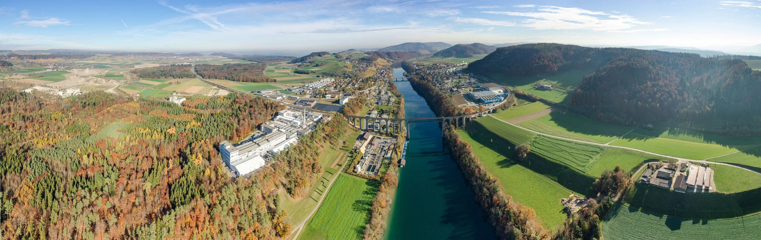 An aerial panoramic view of a river running through a valley, with a dam, industrial buildings, houses, and green fields surrounded by hills with forests.