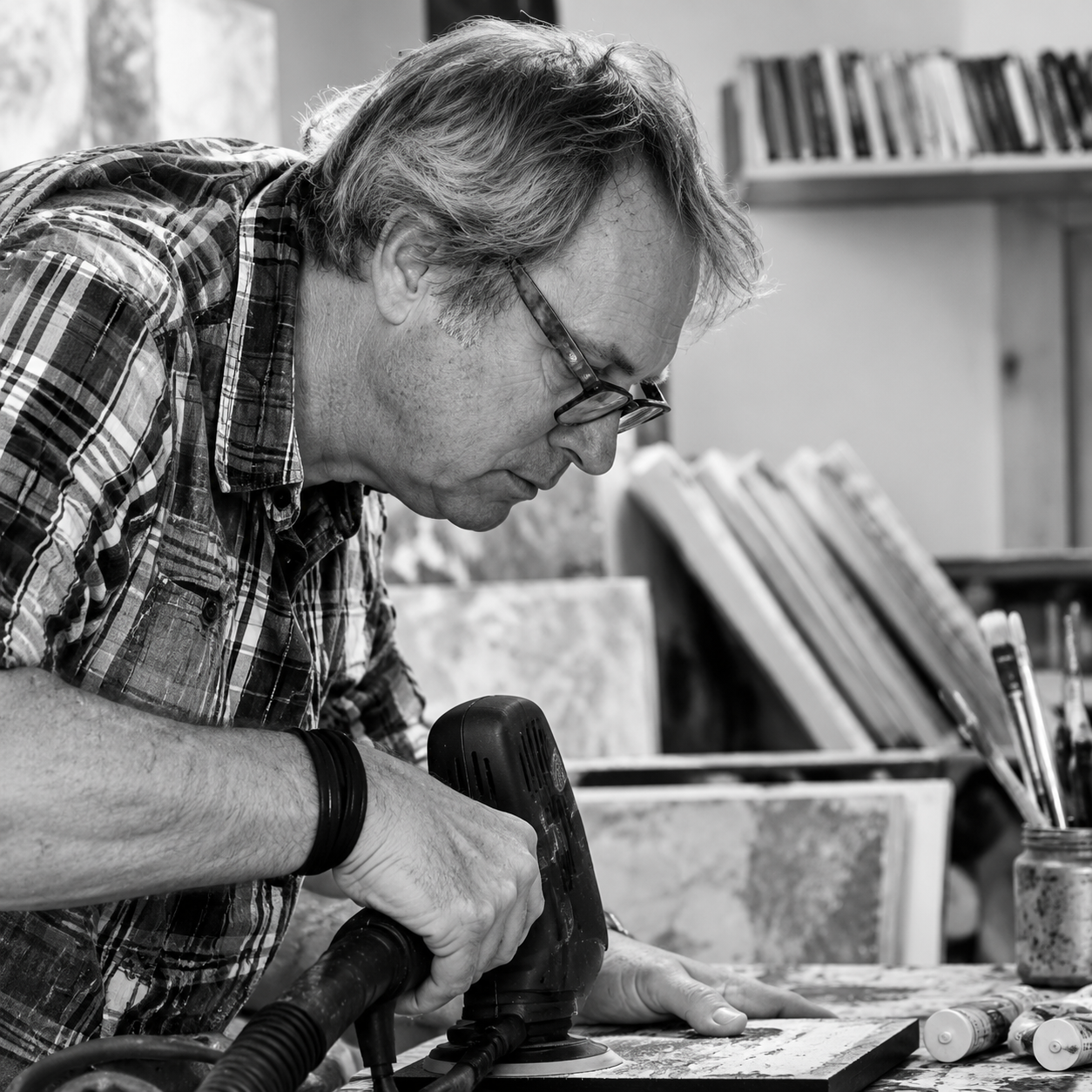 A man wearing glasses and a plaid shirt using a power sander on a piece of wood in a workshop with shelves of books and art supplies in the background.