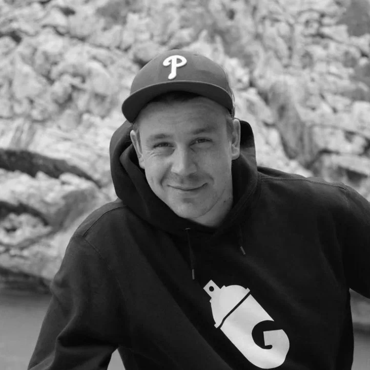 A young man wearing a baseball cap with a 'P' logo, a hoodie, and a sweatshirt with a can opener graphic, outdoors against a rocky background, smiling at the camera.