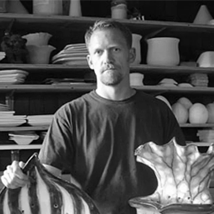 A man with a beard and mustache standing in front of shelves filled with pottery, holding a striped ceramic pumpkin in his left hand and a large ceramic leaf in his right hand.