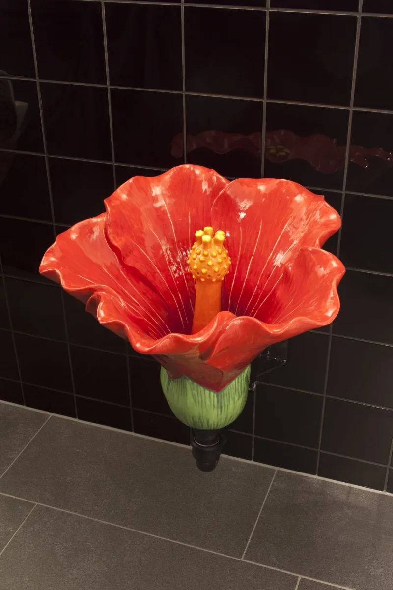 Decorative sculpture of a red flower with a yellow central stamen, mounted on a green and black pipe on a black tiled wall.