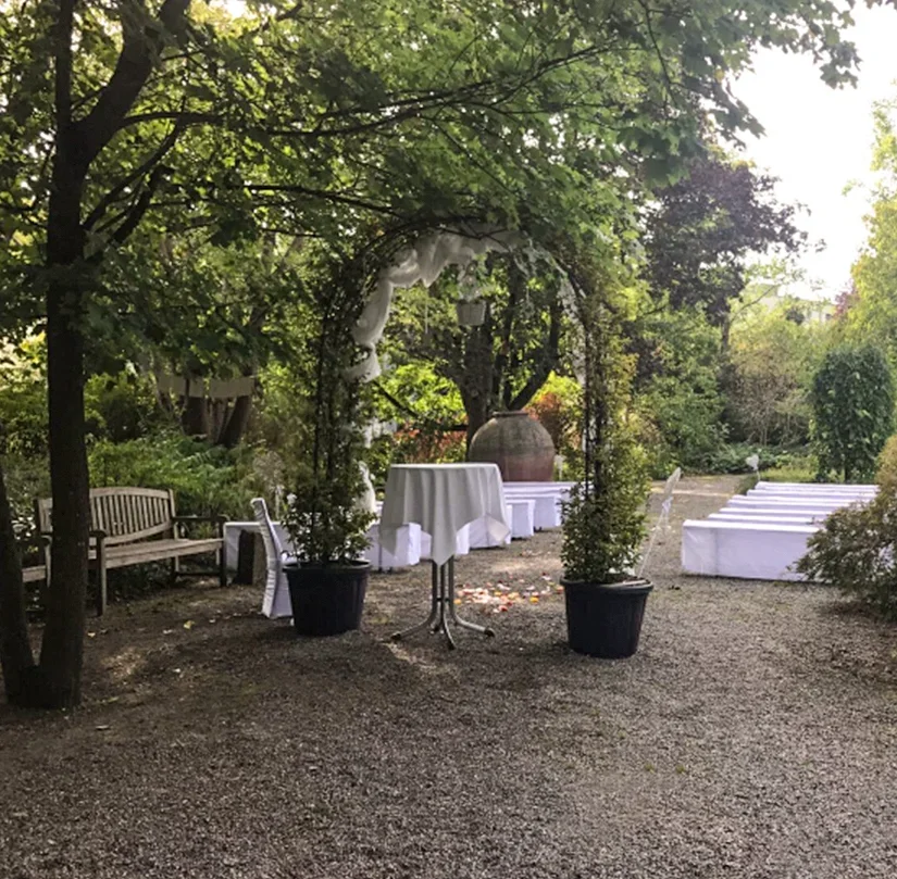 An outdoor garden setup with a white tablecloth-covered table, potted plants, an arbor, a bench, and white draped chairs in the background, surrounded by lush green trees.