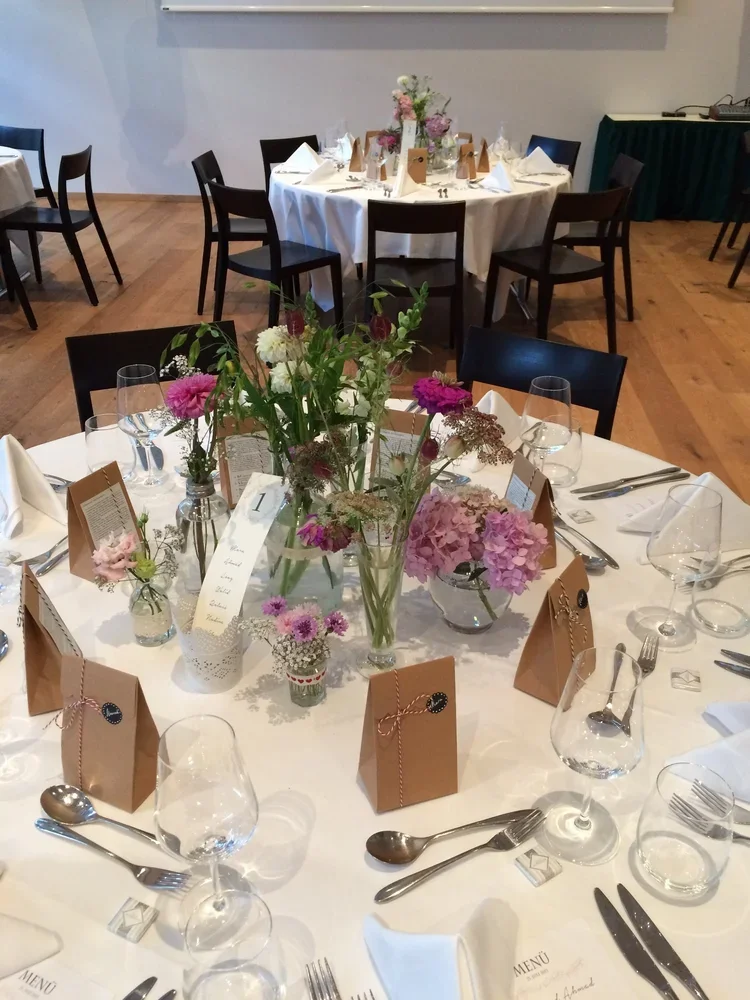 A round banquet table decorated with pink and white floral centerpieces, surrounded by black chairs, set with white napkins, glassware, silverware, and individual brown paper favor bags, in a formal event setting.