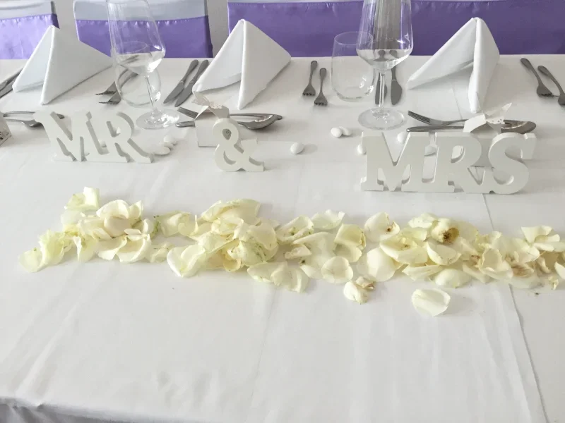 Wedding table decorated with 'Mr & Mrs' signs, white rose petals, white napkins folded in triangles, and glassware.