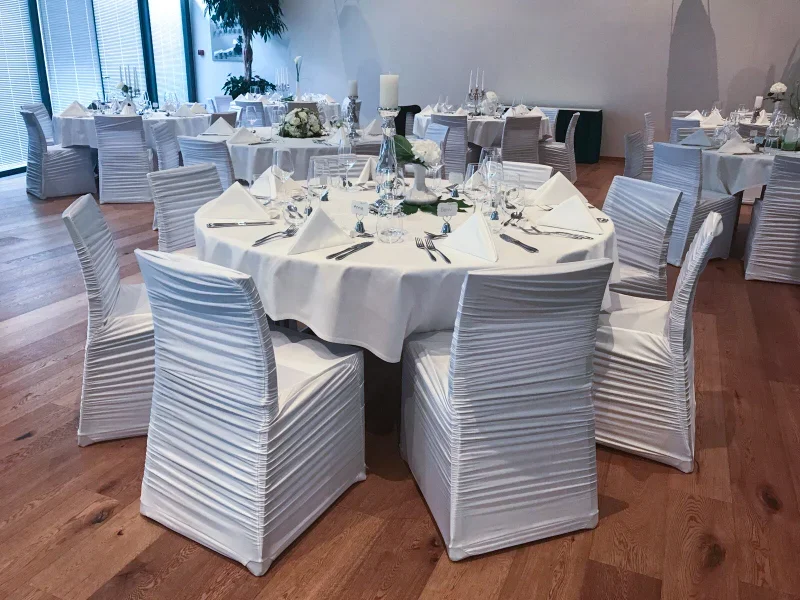 Elegant banquet hall with round tables covered in white tablecloths, set with silverware, wine glasses, and white napkins folded into triangles. White chairs with pleated covers surround each table. The room has wooden floors and large windows with natural light.