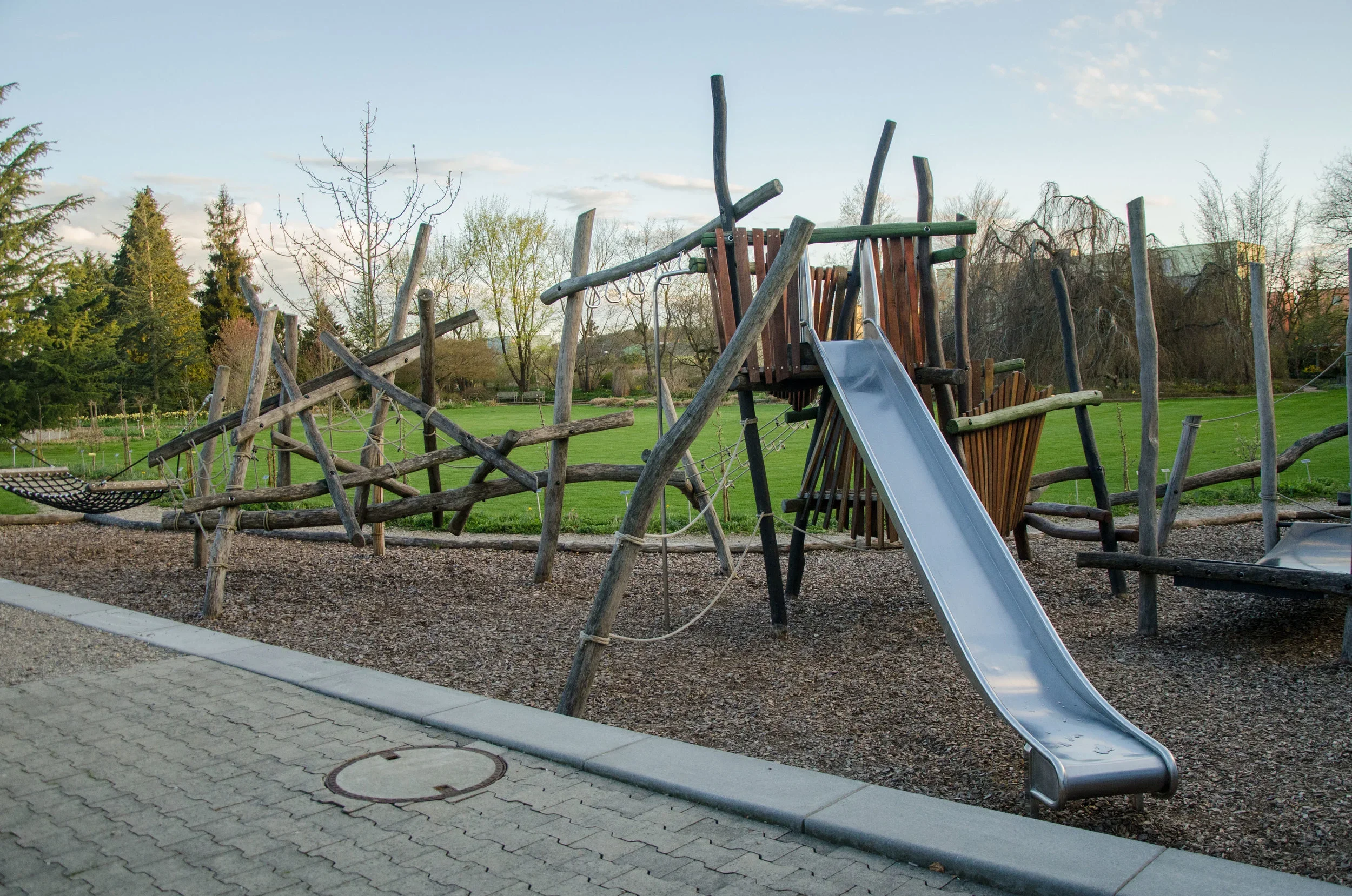 Spielplatz mit Klettergerüst, Rutsche und Wippen im Park, umgeben von Bäumen und Rasen, bei Sonnenuntergang.